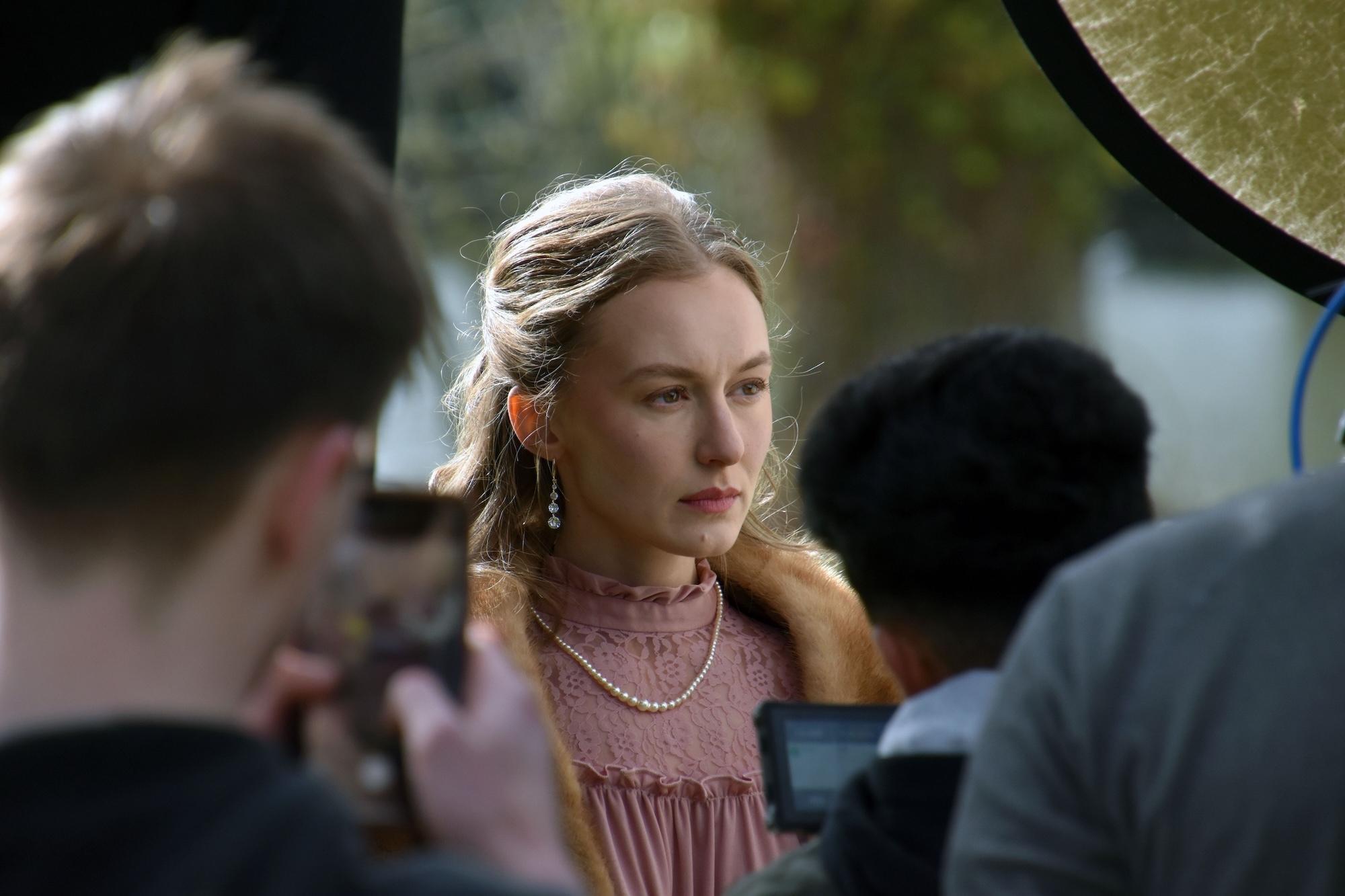 Close-up of actress in pink lace top with layered necklaces surrounded by film crew members and equipment outdoors.