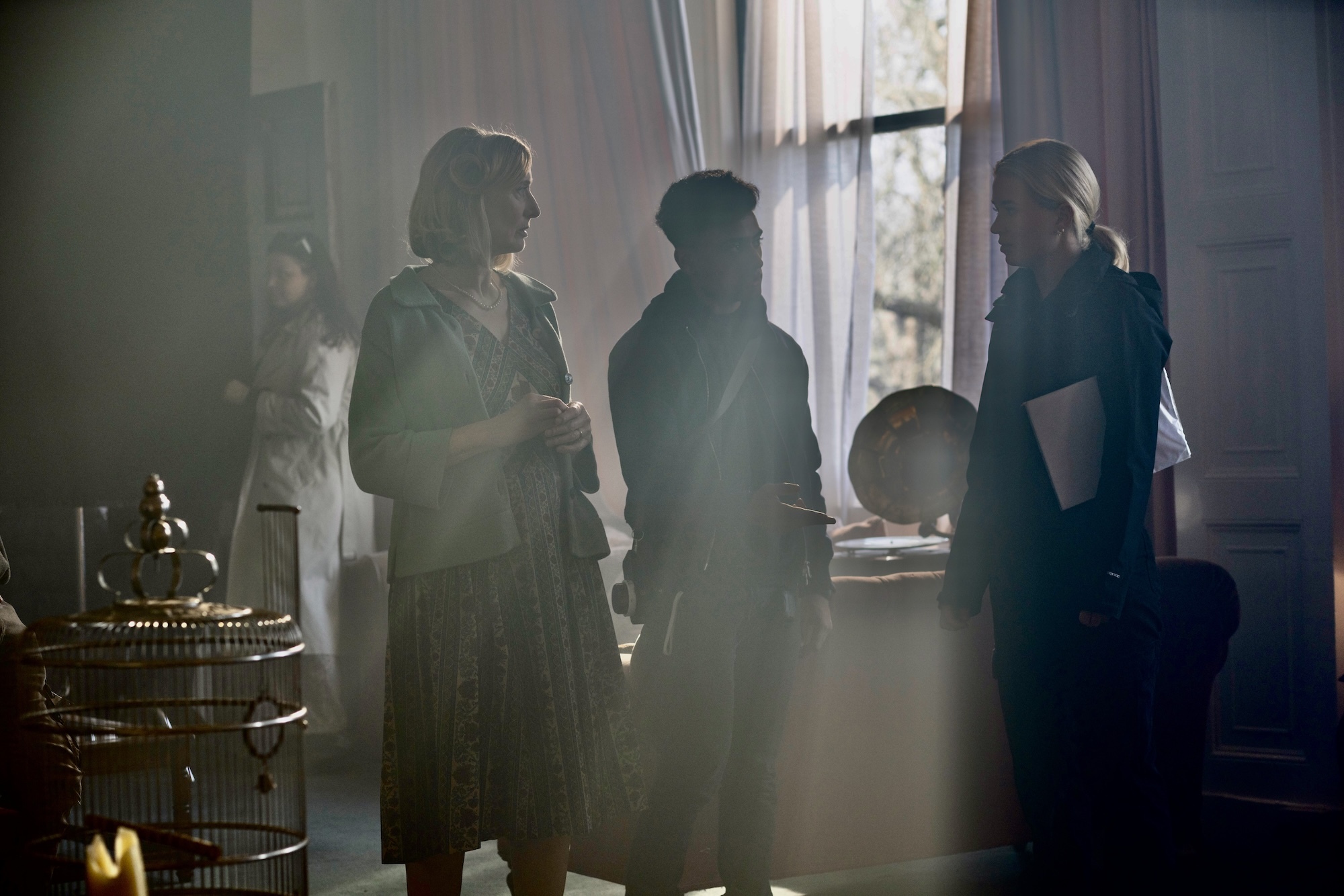Three people standing in dimly lit film set with sunlight streaming through tall window, creating hazy atmospheric lighting.