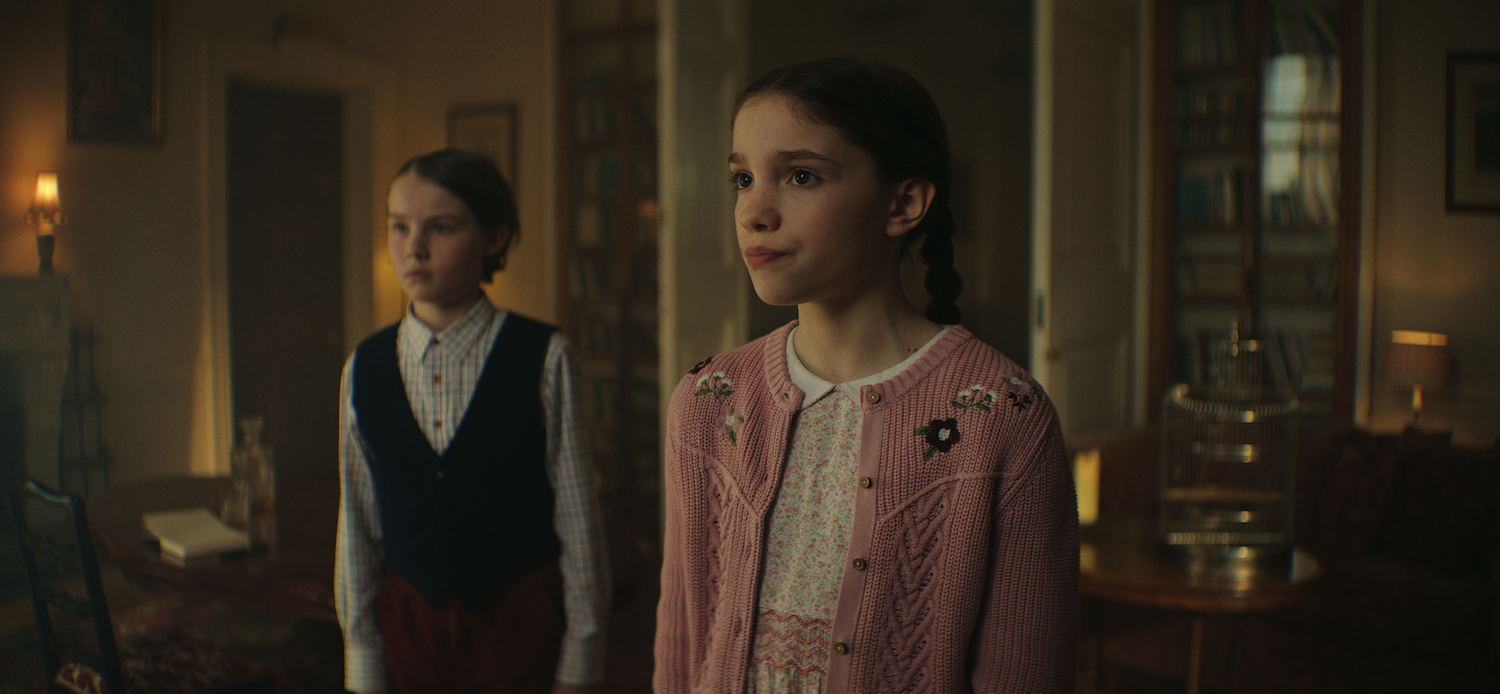 Two young people in period costume stand in dimly lit room with warm lighting. One wears dark waistcoat, other in pink cardigan.