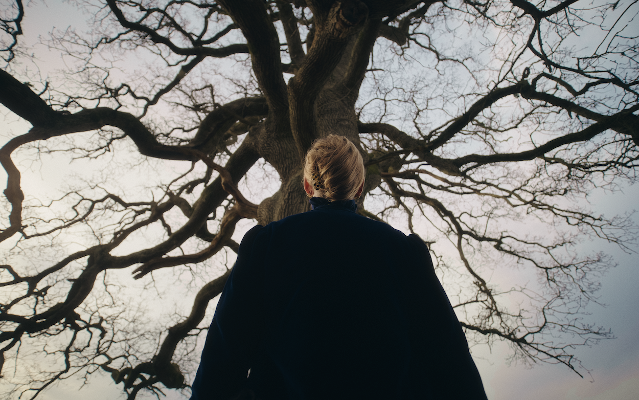 Person in dark coat viewed from below standing beneath bare tree with twisted branches against grey sky.