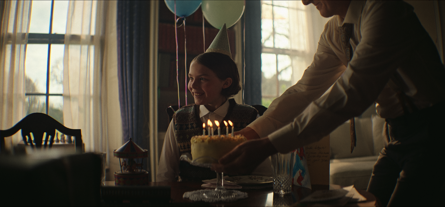 Person sitting beside birthday cake with lit candles as another person reaches towards cake. Balloons and window visible in background.