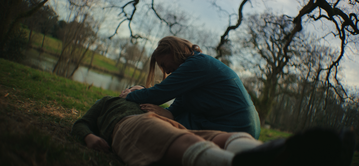 Person in dark teal jacket kneeling beside someone lying on grass under bare winter trees with overcast sky.