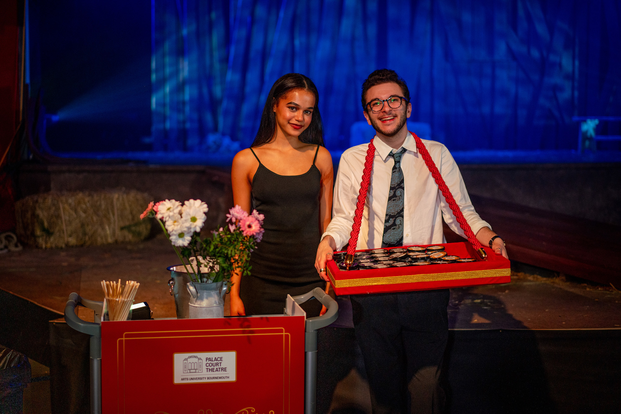 AUB Productions' 'The Ballad of Maria Marten' – two actors off-stage with flowers, one has a red ice cream carrier slung around his neck, blue background,