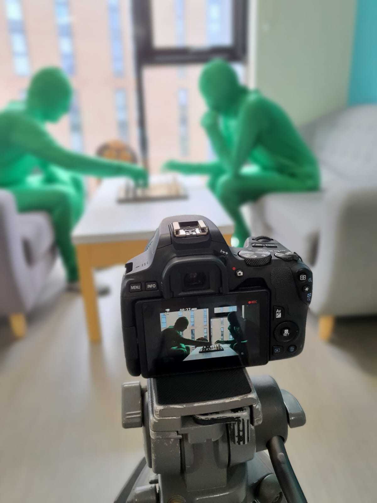 A camera filming two people wearing morph suits and playing chess.