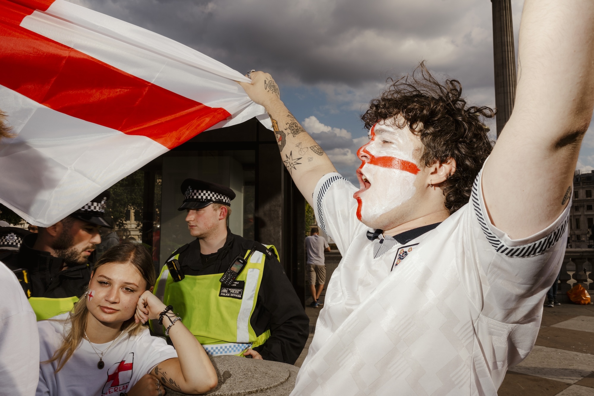A football fan cheering with arms raised holding a large England flag. Next to him, a woman with a bored expression resting her head on her hand, with two police officers behind her.