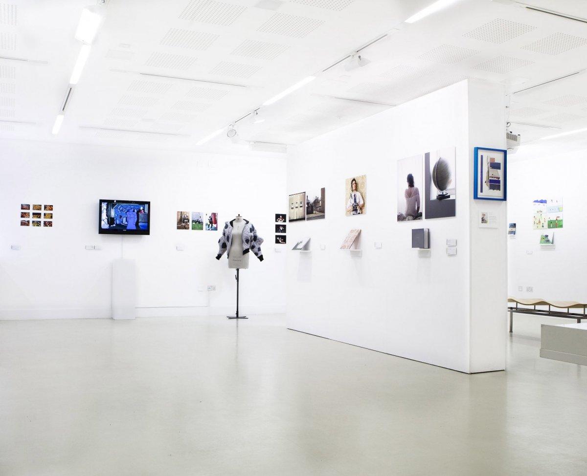 A gallery room with artworks hanging on all walls, as well as a TV screen and a mannequin dressed with a costume.