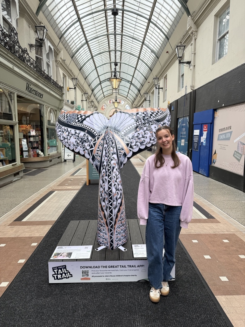 Pia Elliot stood beside her mermaid tail sculpture, floral art deco-style patterns in black, purple and gold, in Westbourne Arcade.