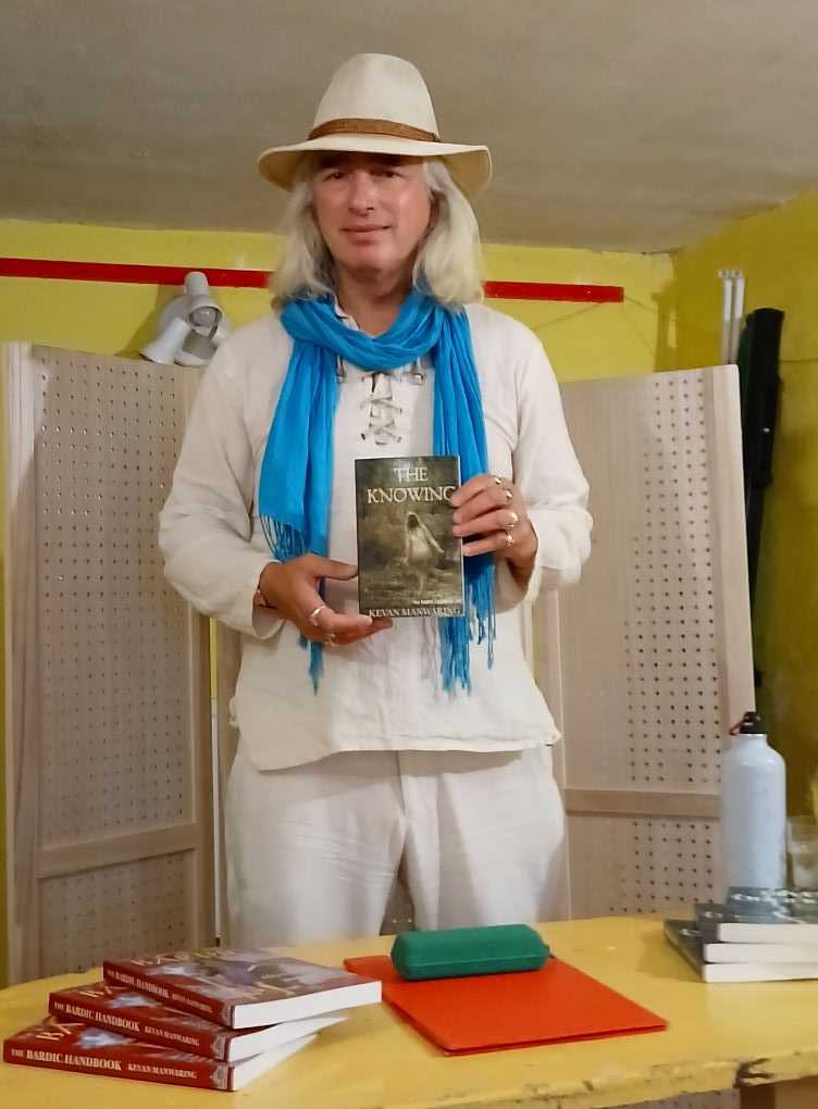 Dr K Manwaring standing behind a desk holding a copy of "The Knowing".