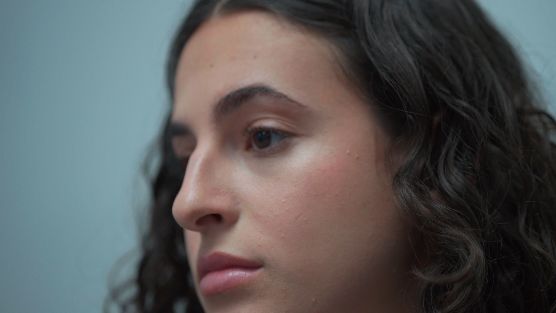 A close-up profile of a young woman with dark curly hair against a pale blue background.