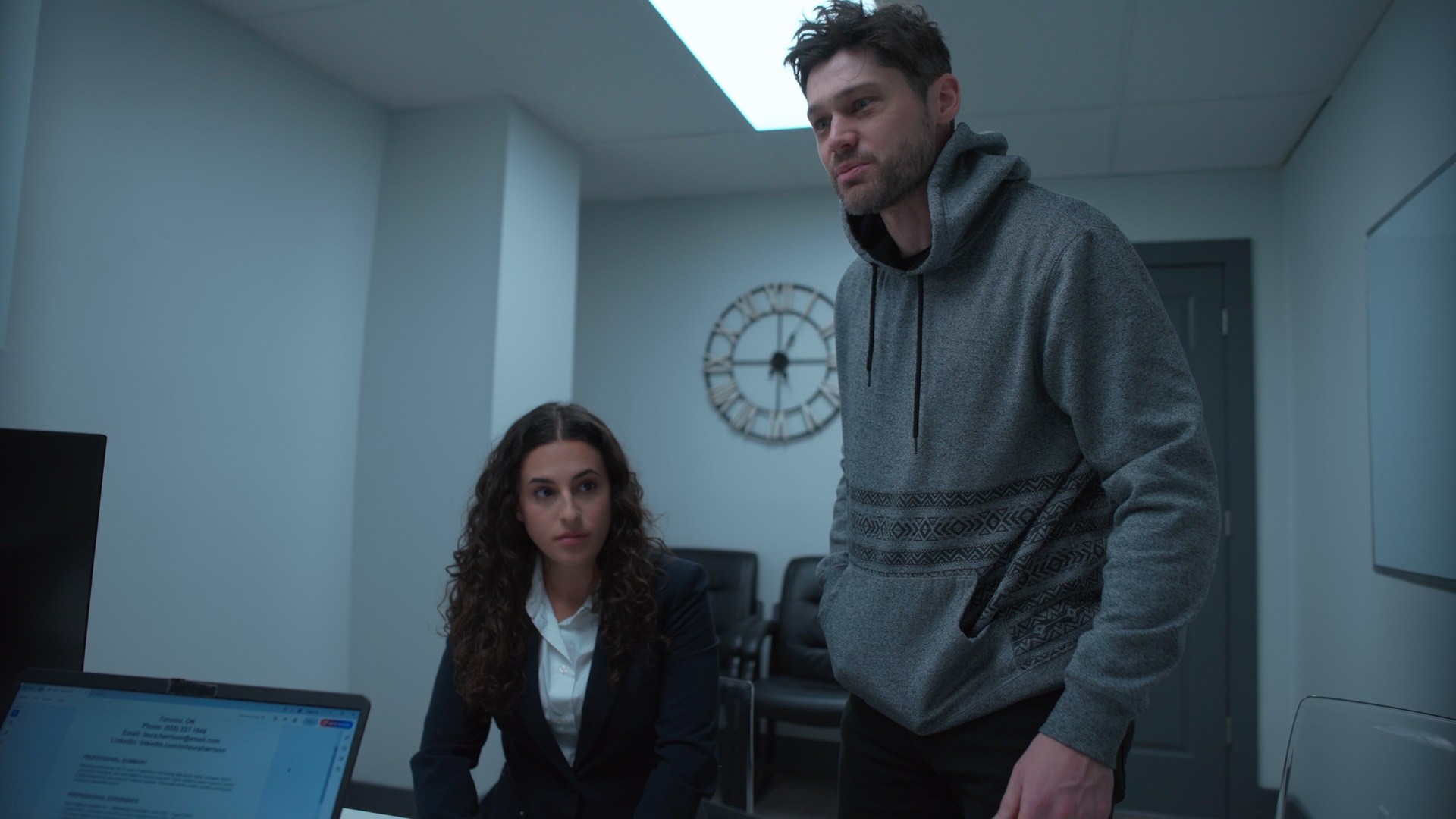 A man in a grey hoodie stands beside a seated woman in a dark blazer in an office, with a wall clock and computer monitor in the background.