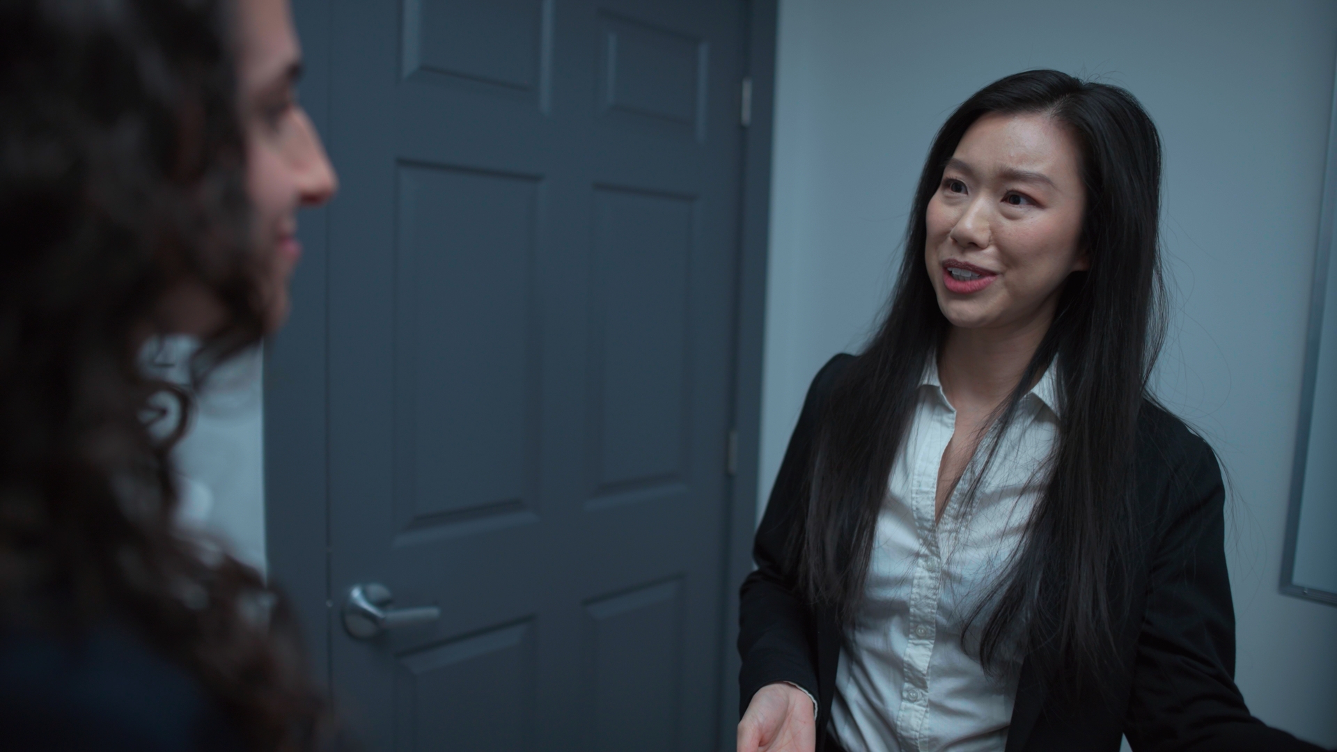 A woman in a black blazer and white blouse stands by a grey door, speaking to a man in the foreground.