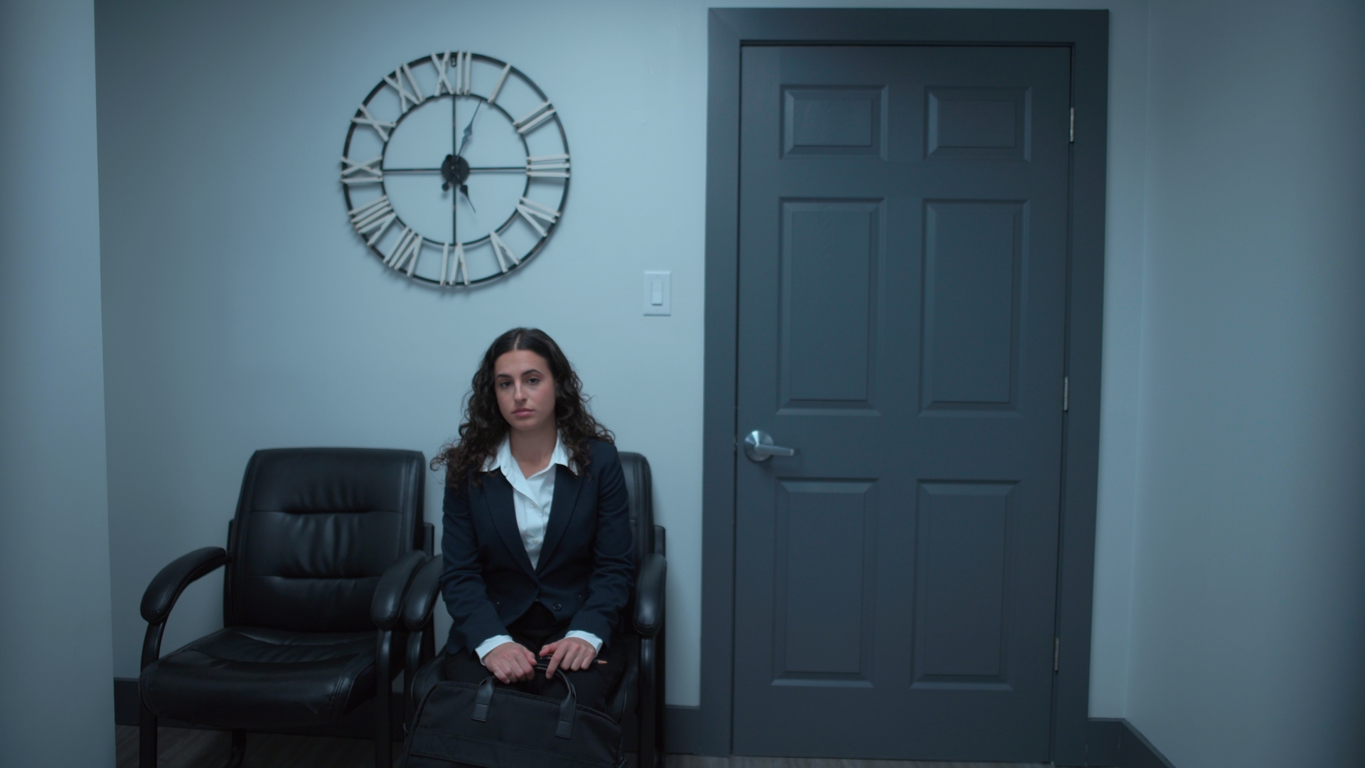 A woman in a dark suit sits against a grey wall beside a black office chair, with a large wall clock above her and a closed grey door to the right.