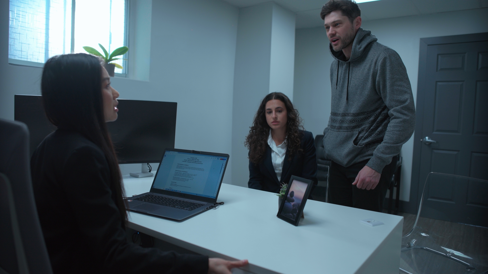 Three people are gathered around a desk in a small office, with a woman seated beside an open laptop as a man stands and another woman sits nearby.
