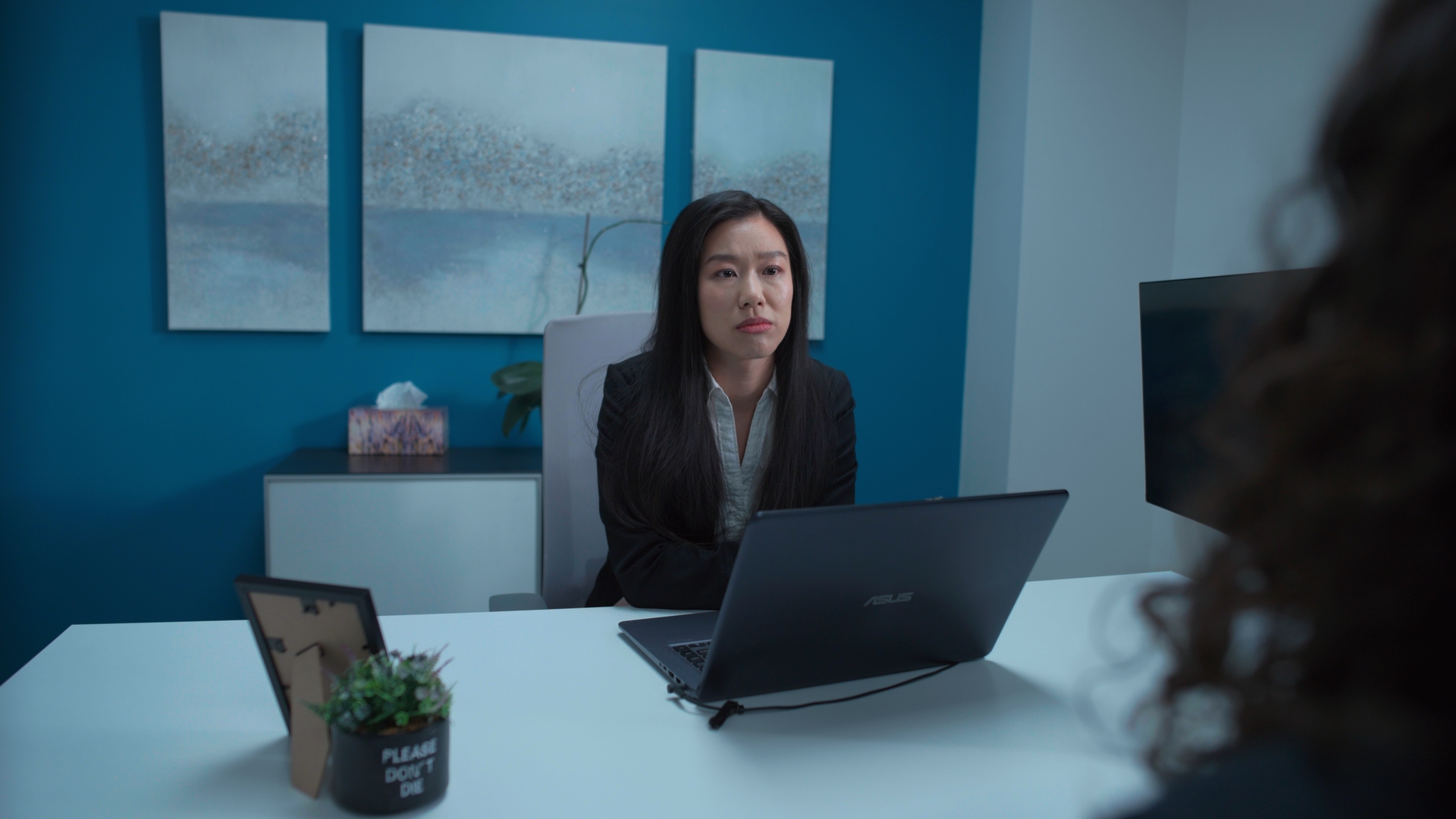 A woman in a dark blazer sits behind an open laptop at a desk in a modern office, with framed artwork on a blue wall and a tissue box on a cabinet behind her.