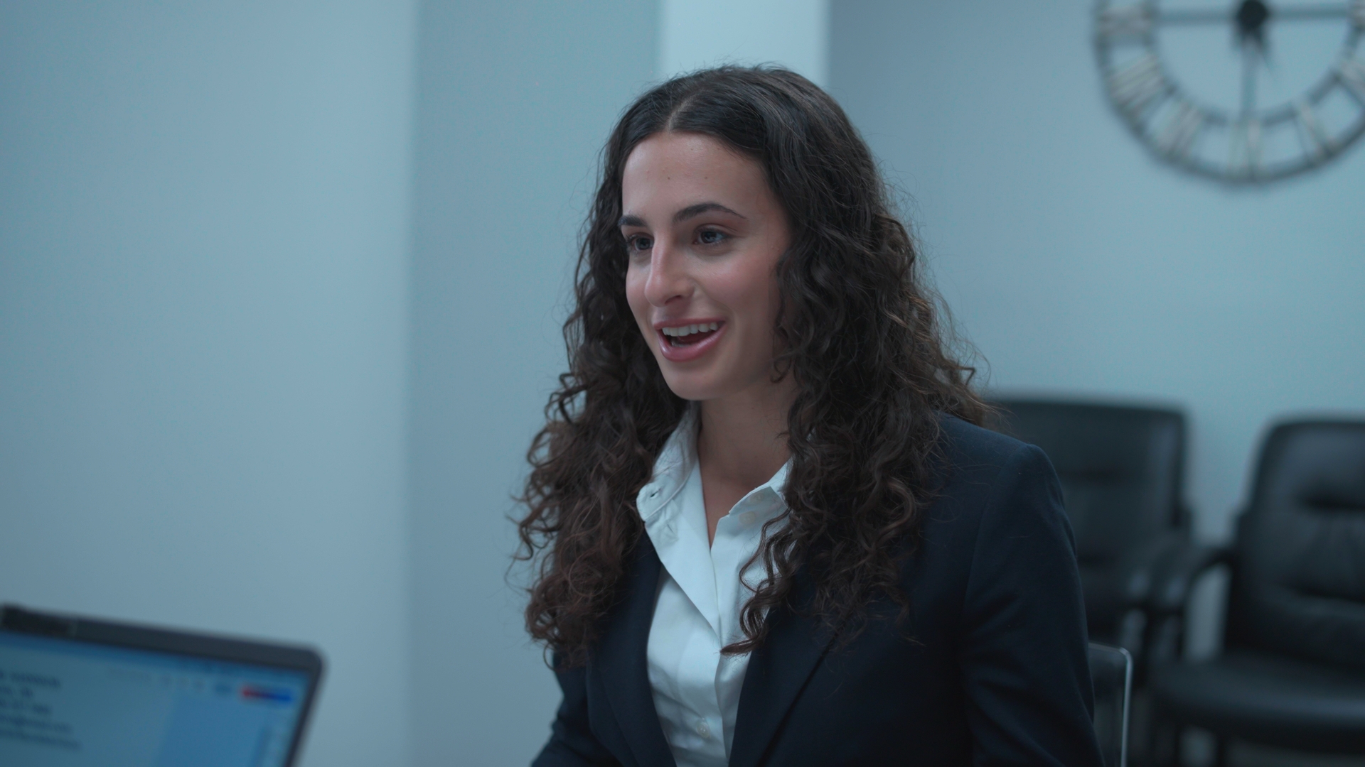 A woman with curly dark hair in a navy blazer and white shirt sits in an office, with a computer screen in the foreground and a wall clock behind her.