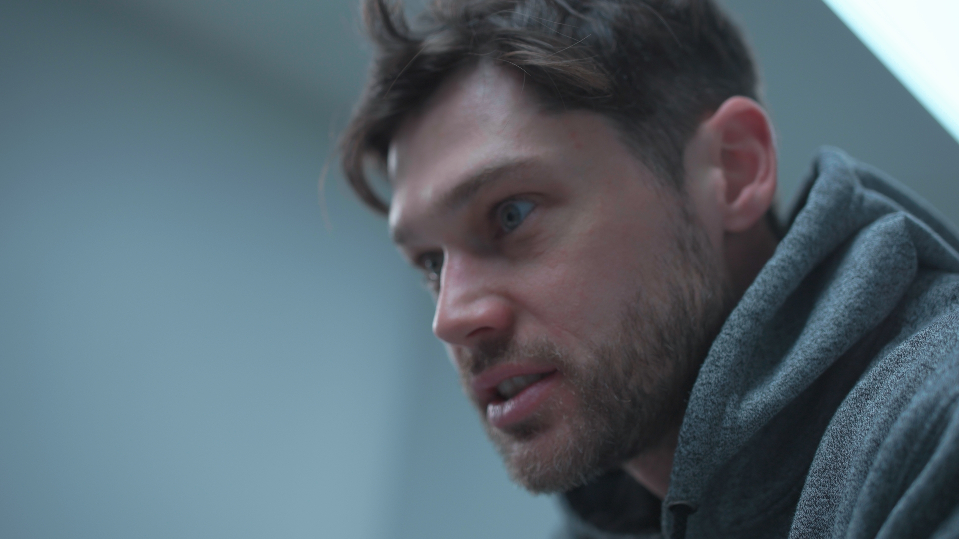 A close-up of a man in a grey hoodie, with a blurred light grey indoor background.