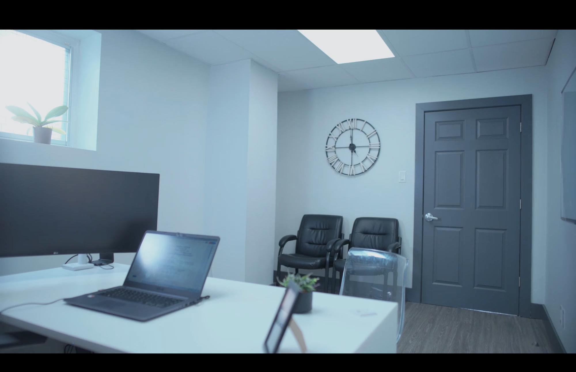A modern office with a laptop on a white desk in the foreground, black chairs against the back wall, and a grey door beneath a wall clock.