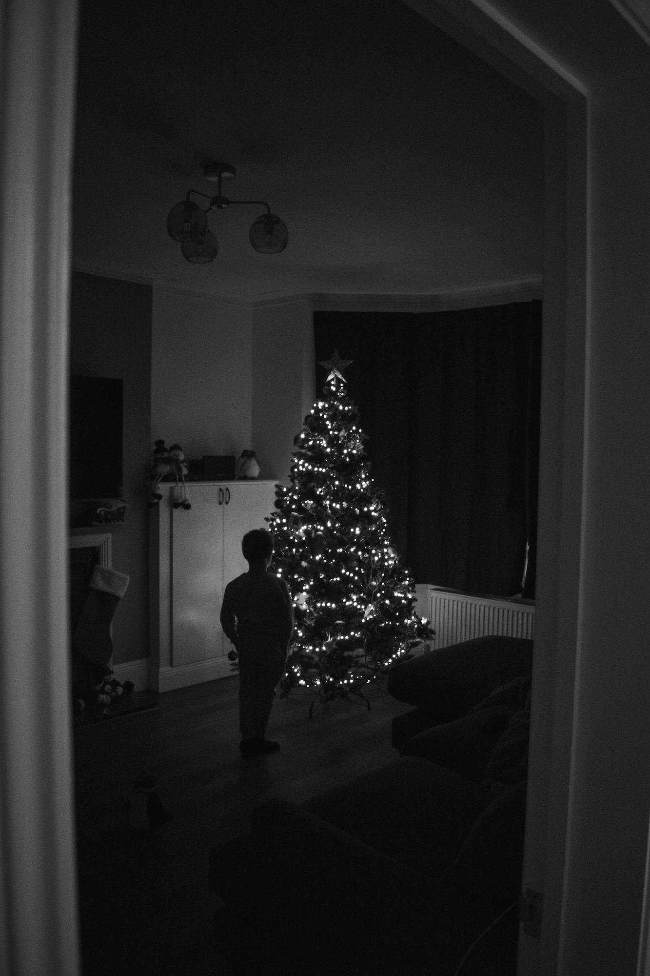 A child stands in a dimly lit living room beside a lit Christmas tree, with a white cabinet and dark curtains in the background.