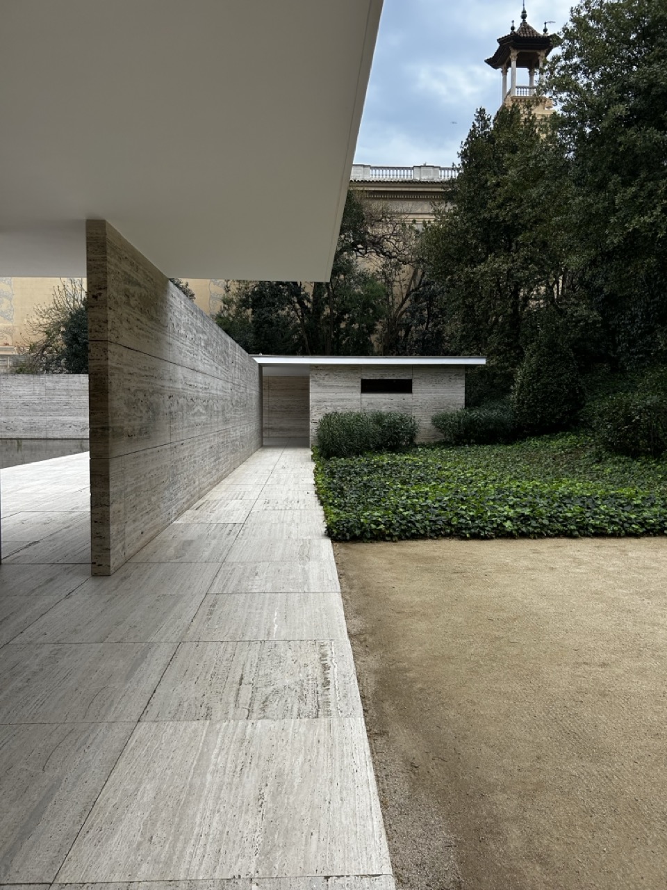The Meis van de Rohe pavilion, large concrete floor, wall and structure to the left, concrete building at the end, trees and foliage to the right.