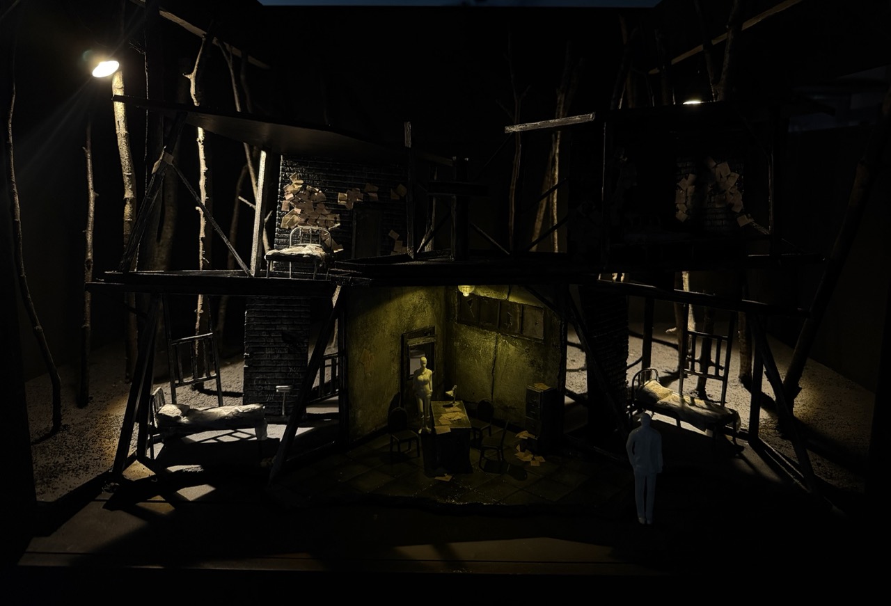 A dark theatre stage shows a damaged multi-storey set with ladder-like beams, lit by a single overhead lamp and a small central room below.