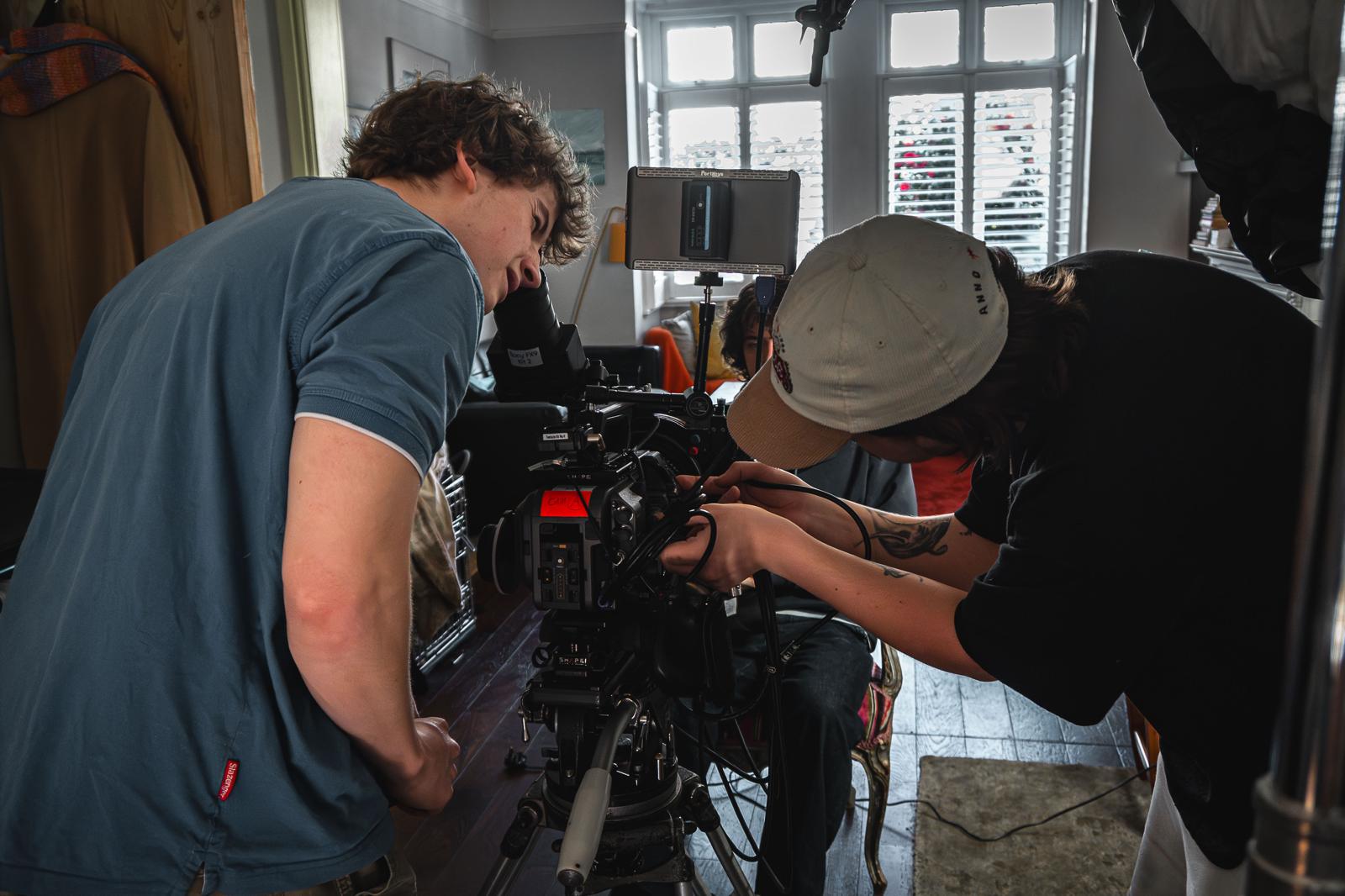 Two individuals are focused on setting up a professional video camera on a tripod inside a room. One person, wearing a blue shirt, leans over to adjust the camera while the other, in a cap, assists with the equipment.