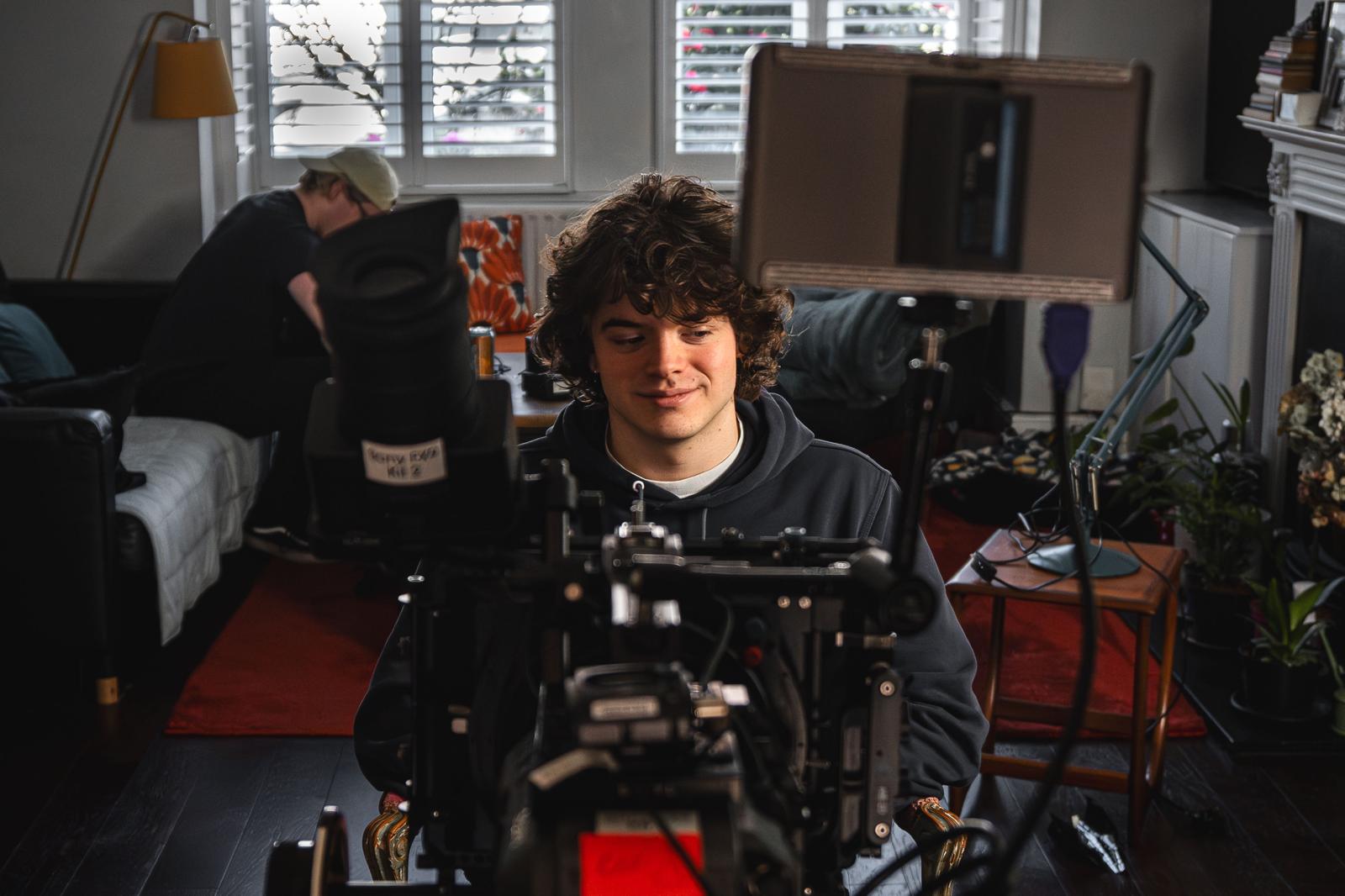 A young man with curly hair sits in front of a professional film camera within a living room setting. In the background, another person is visible engaged in an activity, surrounded by warm lighting and a mix of furniture.