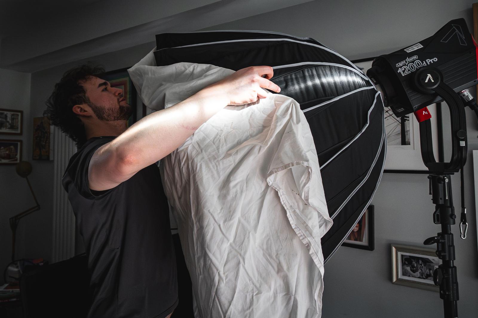 A man with curly hair, wearing a black t-shirt, is adjusting a large black and white softbox light. The room contains several framed photographs on the walls, and a tripod-mounted light is visible in the background.