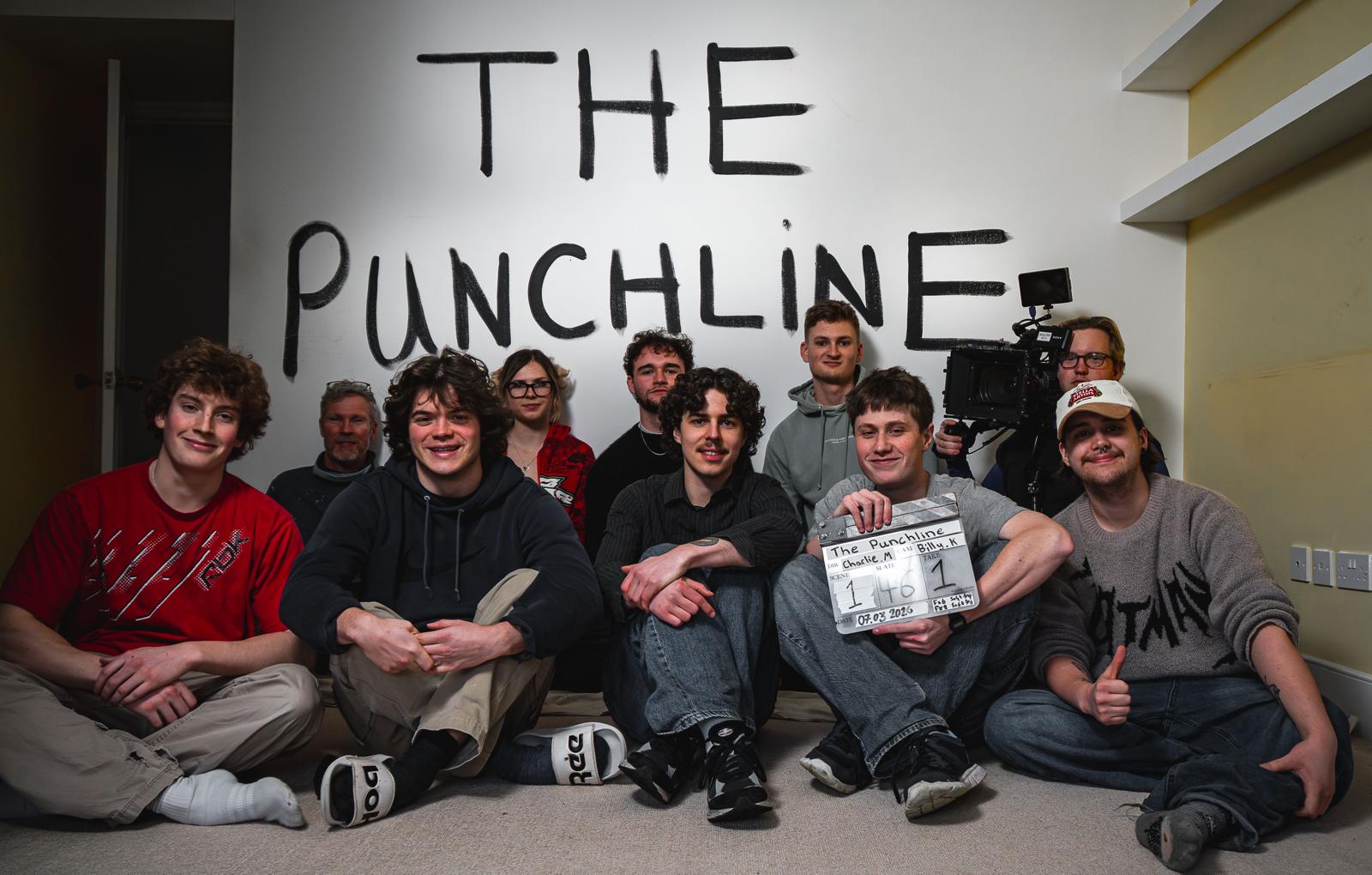 A group of eight individuals sitting on the floor in front of a wall with the text "THE PUNCHLINE" painted in bold letters. One person holds a film clapperboard, and a camera is set up in the background.