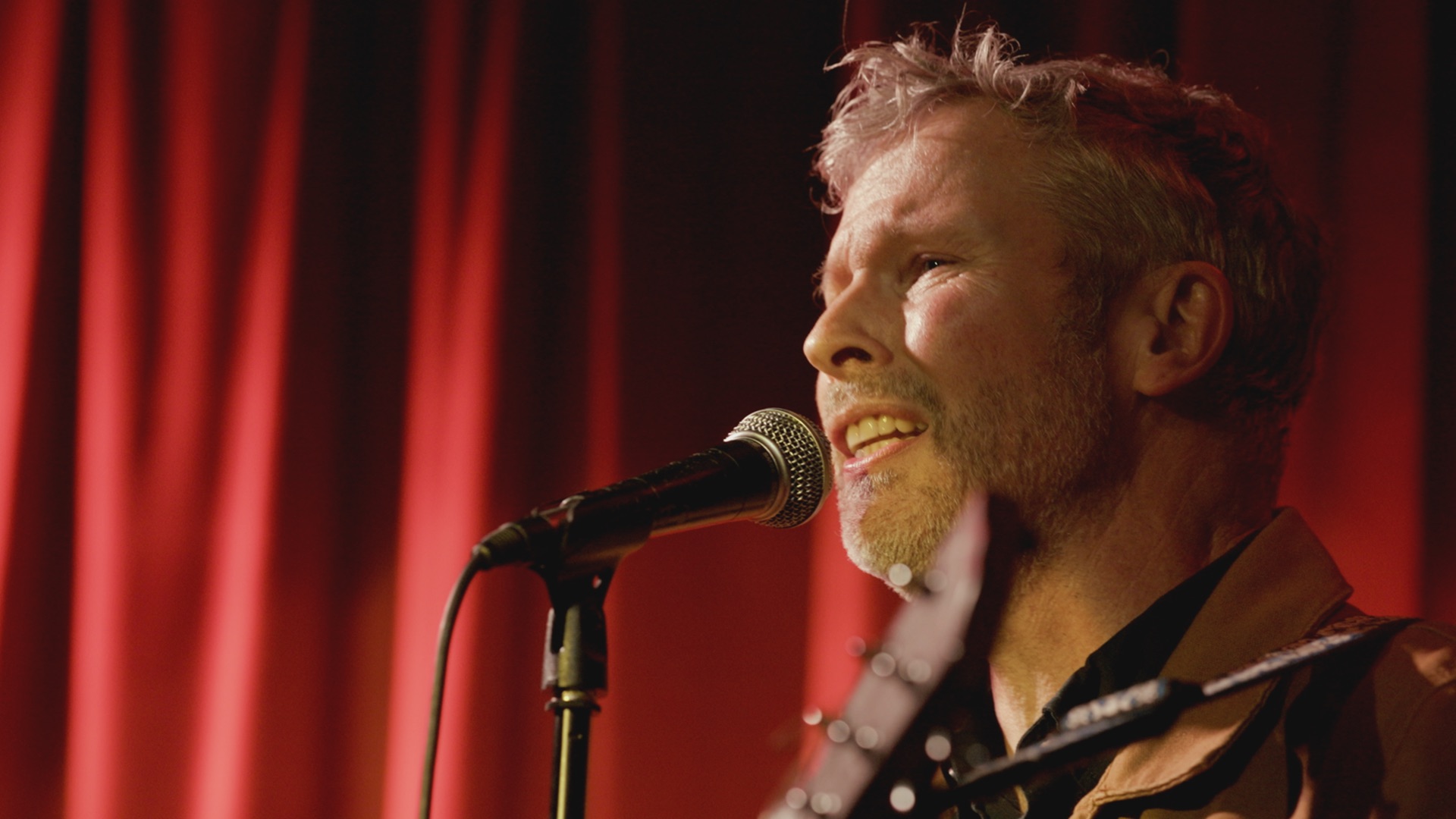 A man with short gray hair singing into a microphone, positioned in front of bright red curtains. He is wearing a brown jacket and there is an acoustic guitar visible over his shoulder.