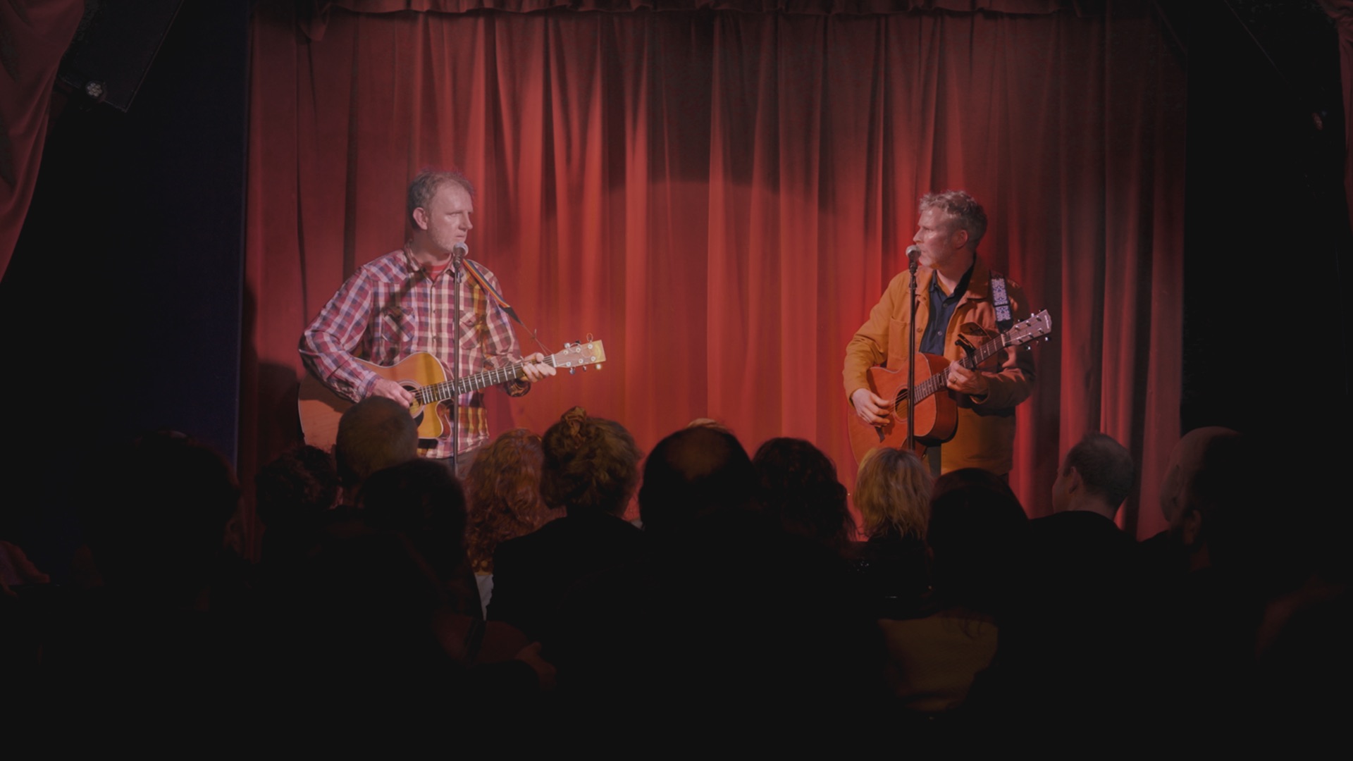 Two musicians perform on stage, one with a plaid shirt and acoustic guitar, the other wearing an orange jacket and playing a smaller guitar. The backdrop is a red curtain, and an audience can be seen in silhouette, listening attentively.