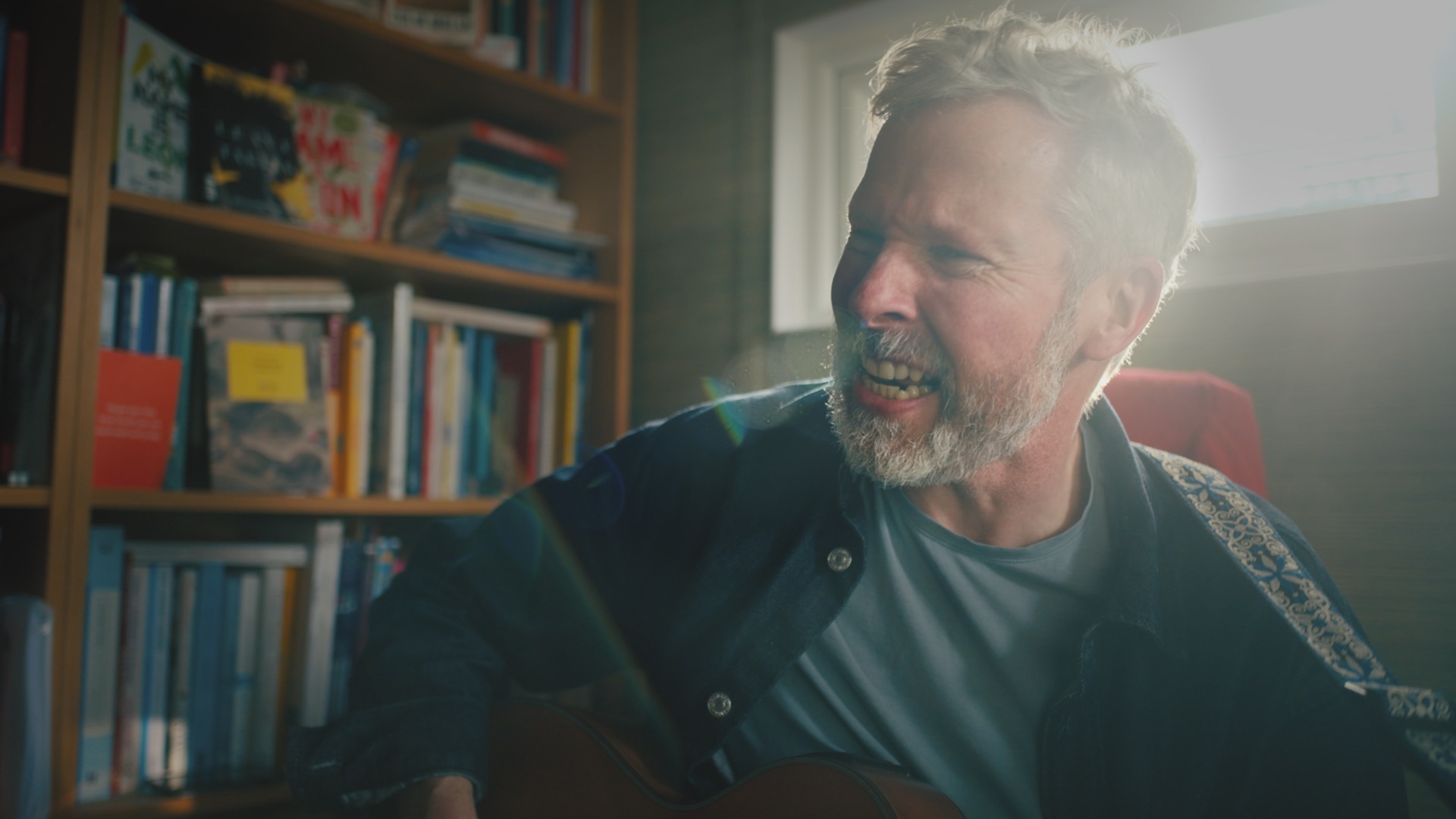 A man with a beard plays acoustic guitar while sitting in a room filled with bookshelves. The background features a window with bright light and various books stacked on the shelves.