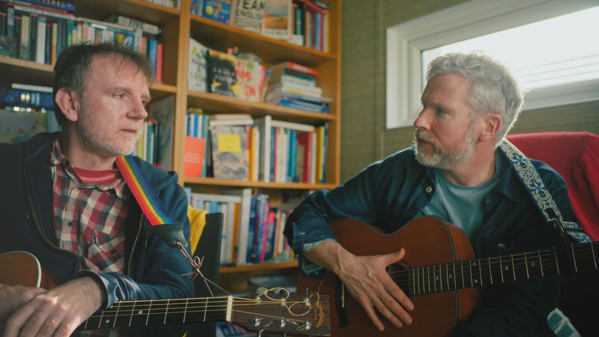 Two men sitting in a room with bookshelves filled with books behind them. One man, in a checked shirt, plays an acoustic guitar while the other, in a blue shirt, holds a guitar and appears to be engaged in conversation.