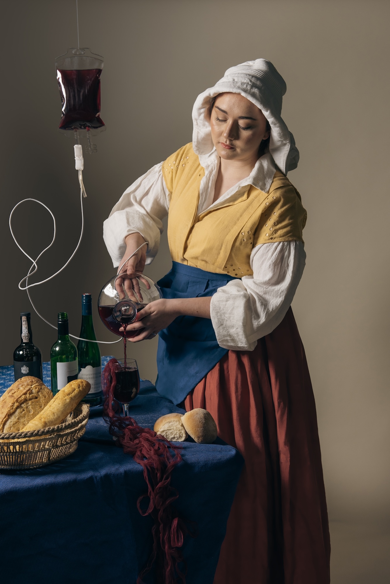 A model dressed like a milkmaid from Vermeer's painting pouring wine into a glass on a table. A basket of bread, bottles of wine and an IV drip with more wine are also on the table.