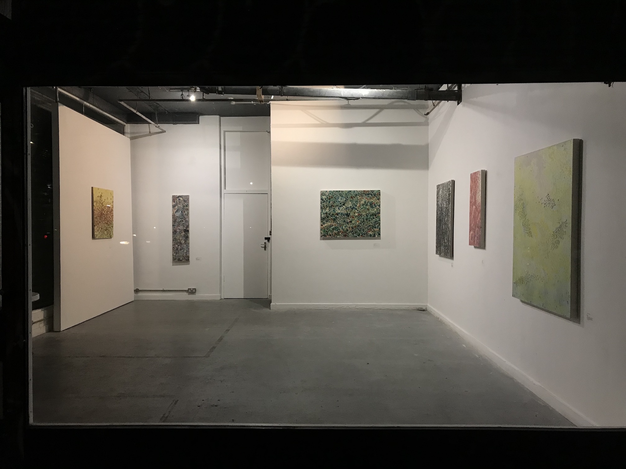 Gallery space with white walls displaying various artworks including green, yellow, and red paintings. Grey floor, exposed ceiling, glass door.