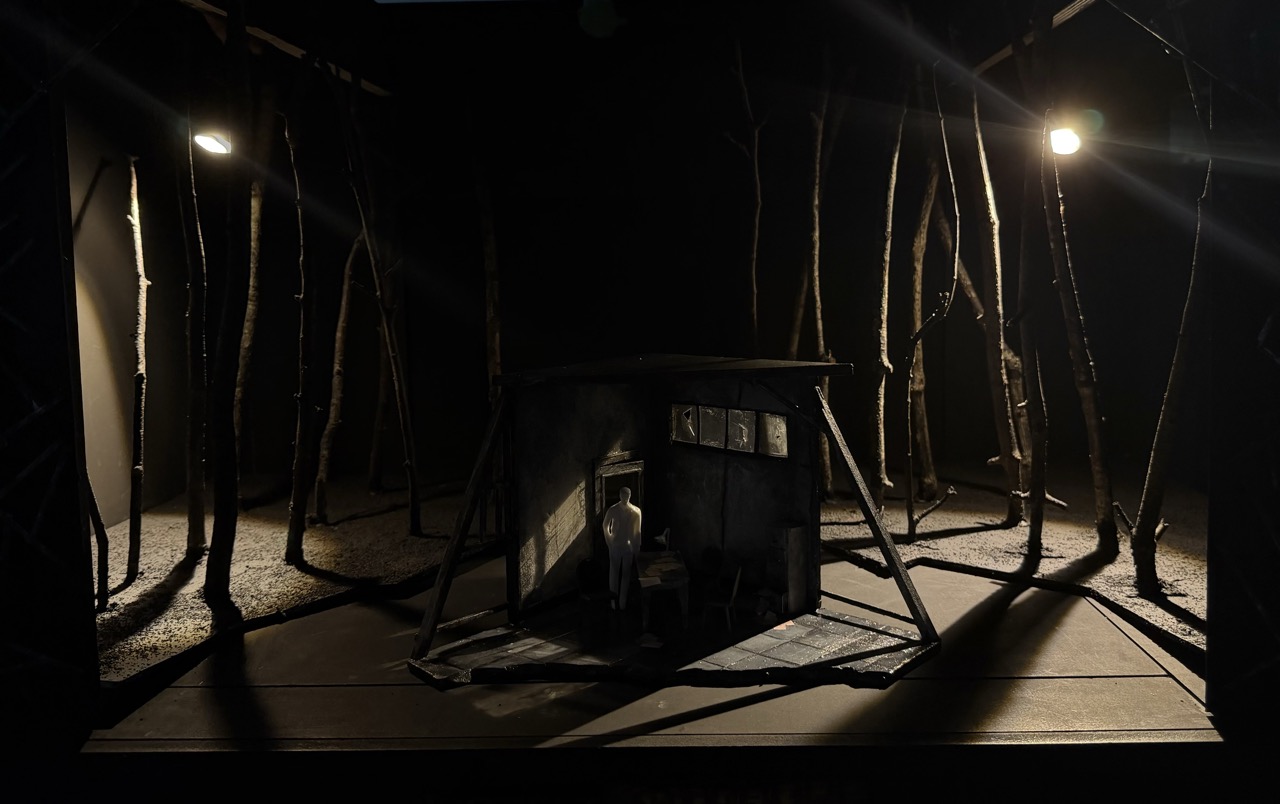 A small dark wooden hut stands on a stage-like platform among tall bare tree trunks, lit by two spotlights in a forest scene.