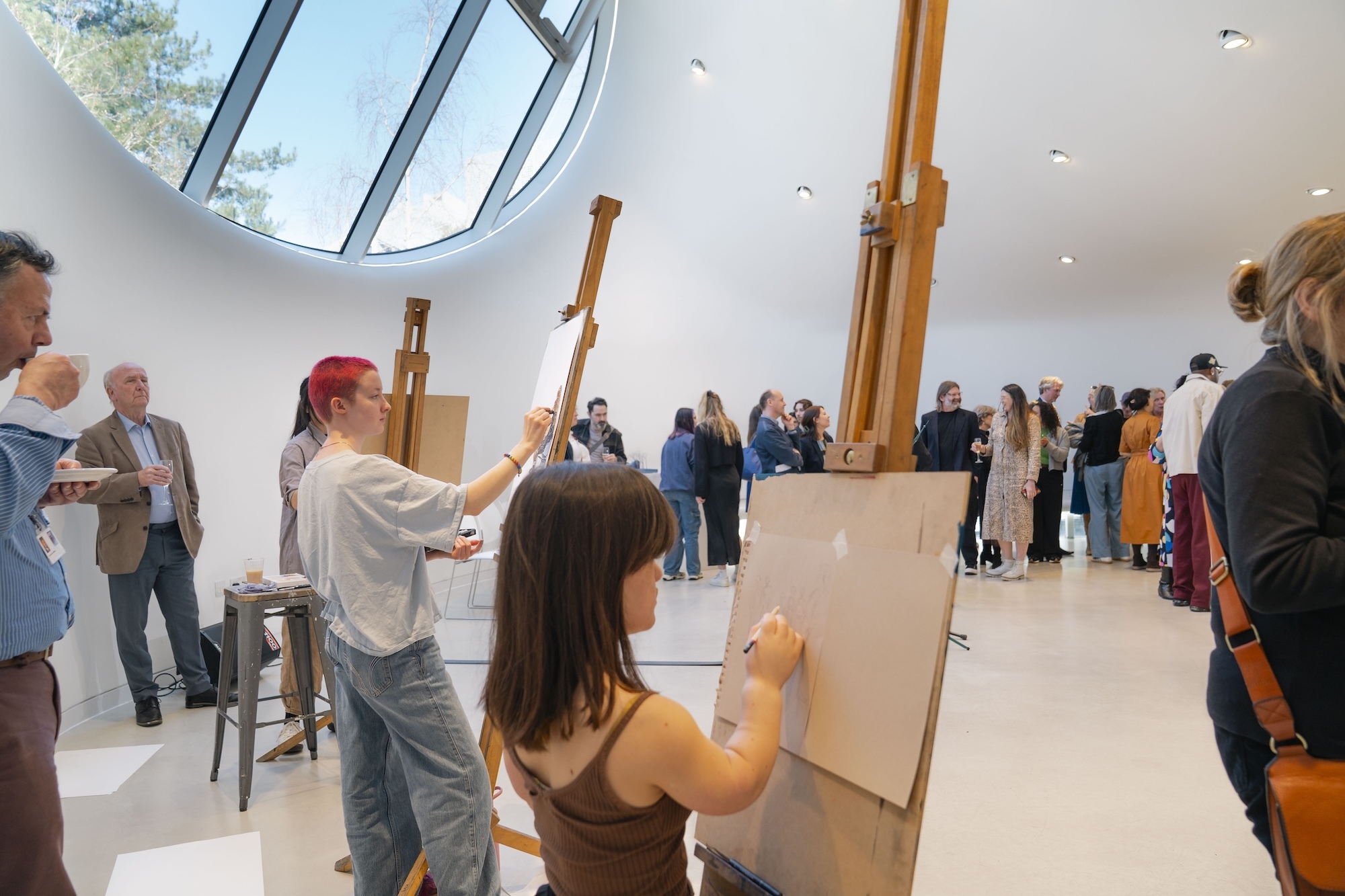 People painting at easels in white gallery space with circular skylight and curved architecture, visitors observing in background.