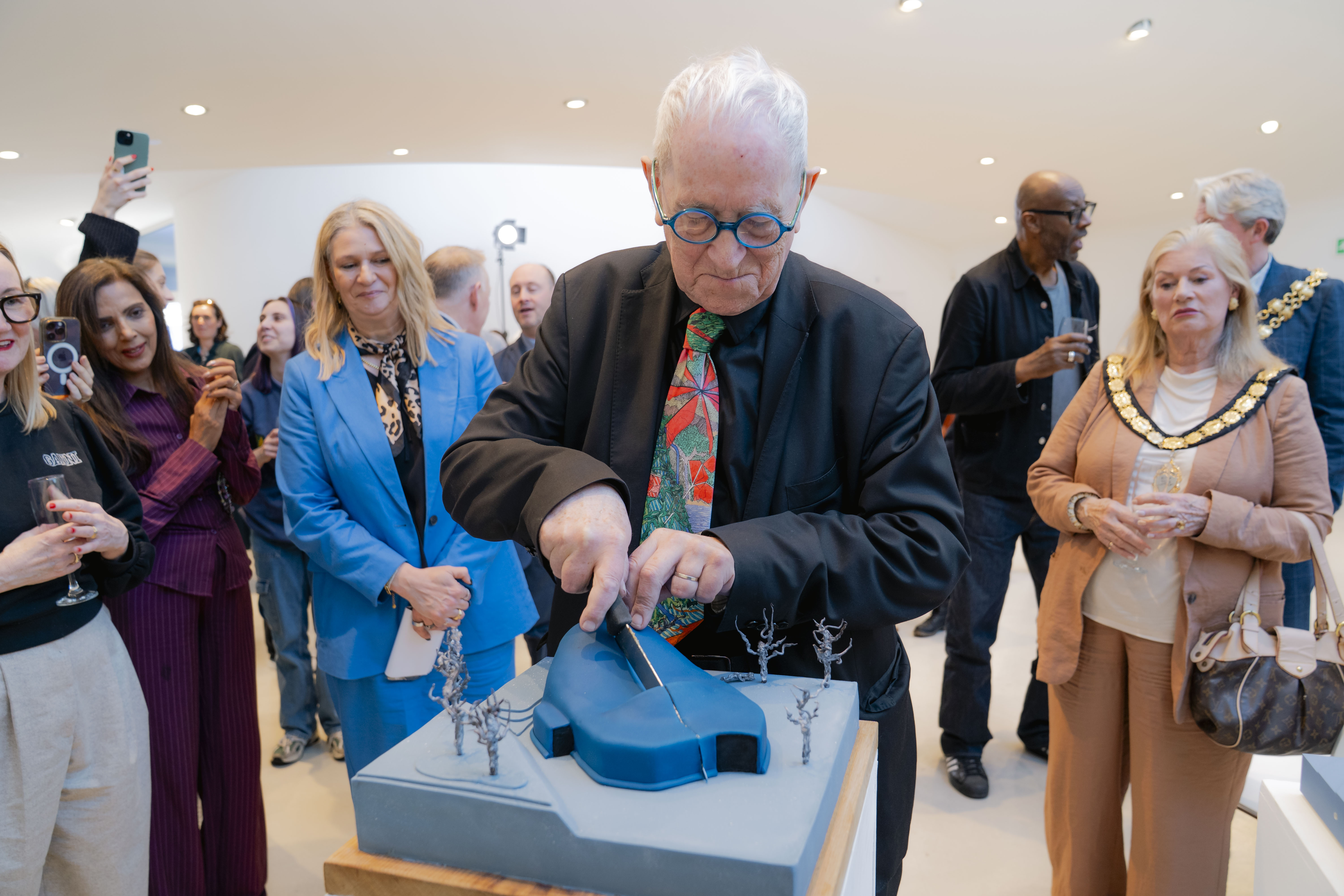 Professor Sir Peter Cook RA cutting into the blue architectural cake model. He has white hair, round blue glasses and colourful tie, surrounded by other people.