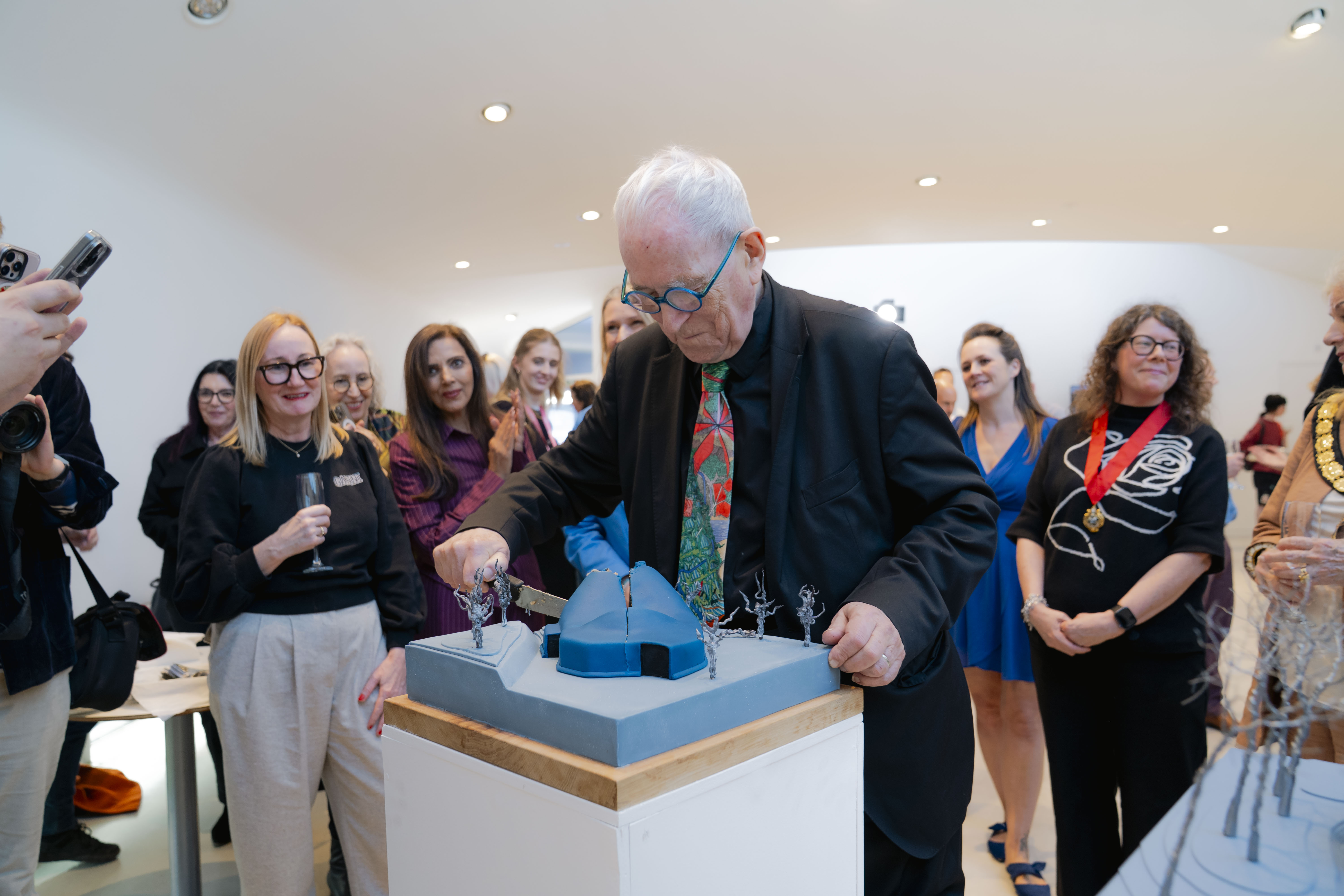 Professor Sir Peter Cook RA cutting into the blue architectural cake model. He has white hair, round blue glasses and colourful tie, surrounded by other people. Side angle.