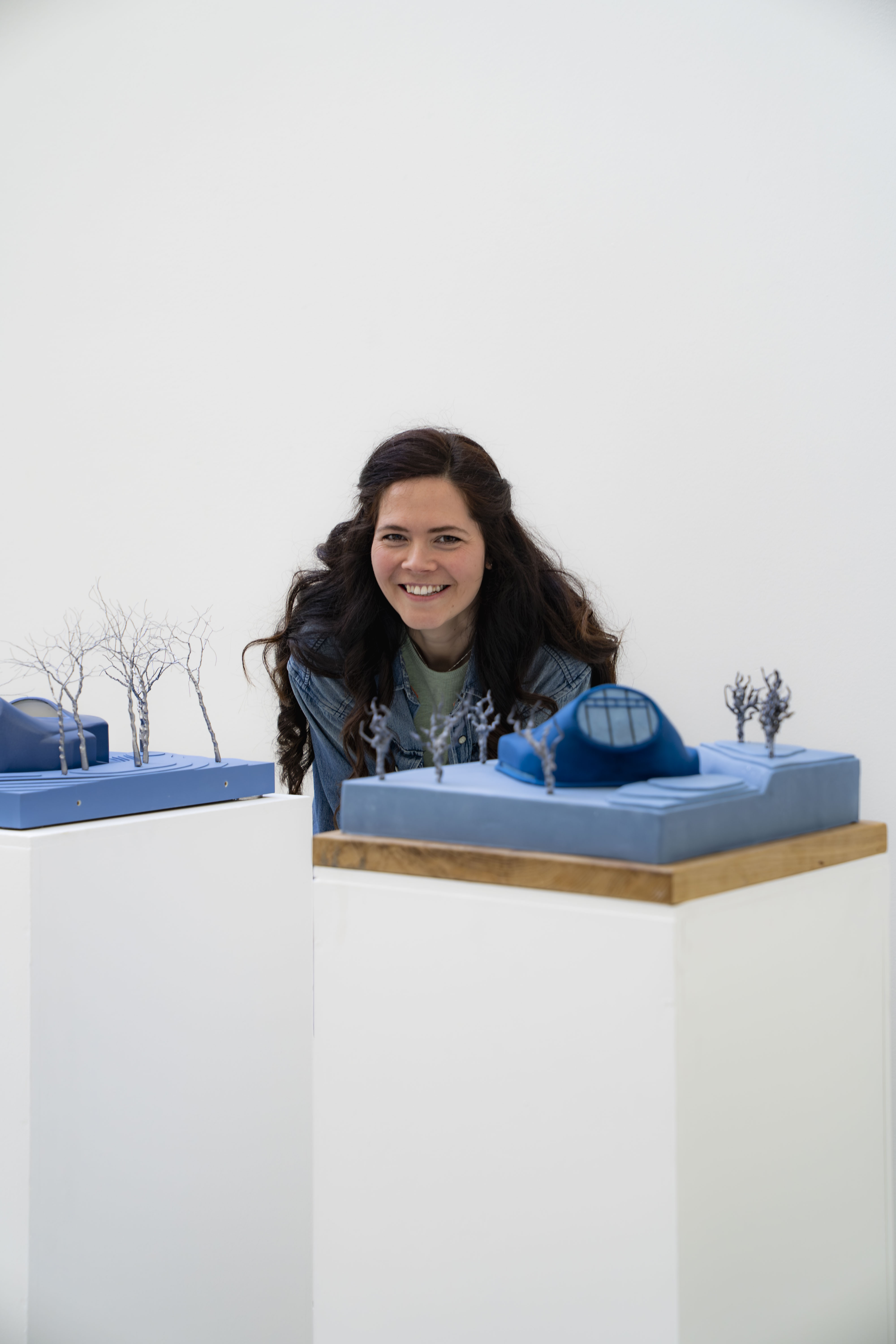 Tasha Diamond stood behind a blue architectural cake and the same model, not cake. She has long dark hair and is smiling, white background.
