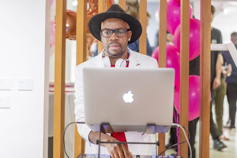 A DJ sitting behind an Apple Mac laptop. There are party balloons and people visible behind them.