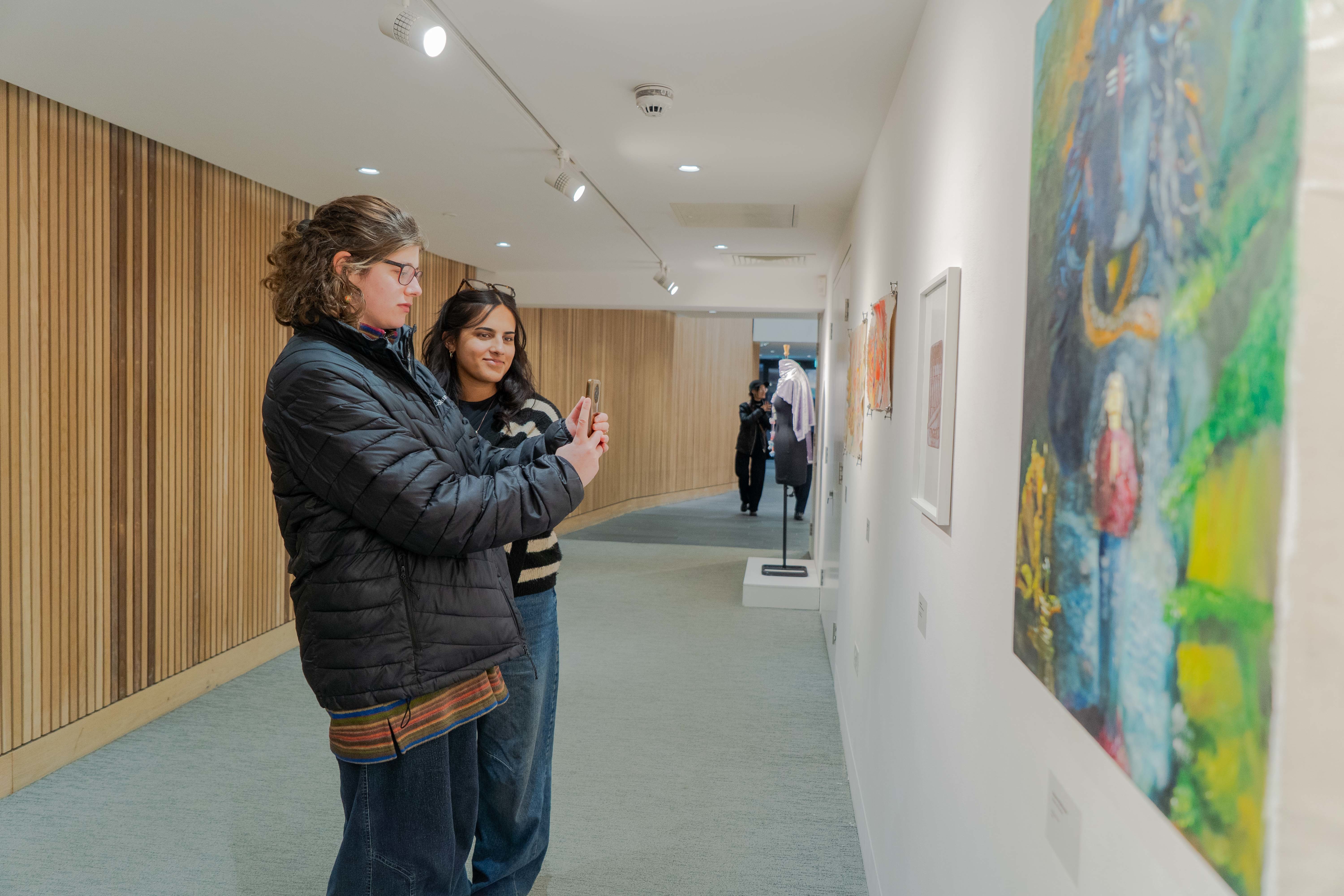 Two visitors to TheGallery during the AUB International Collective 2025 exhibition. One of the visitors is taking a picture of a piece of artwork on their phone.