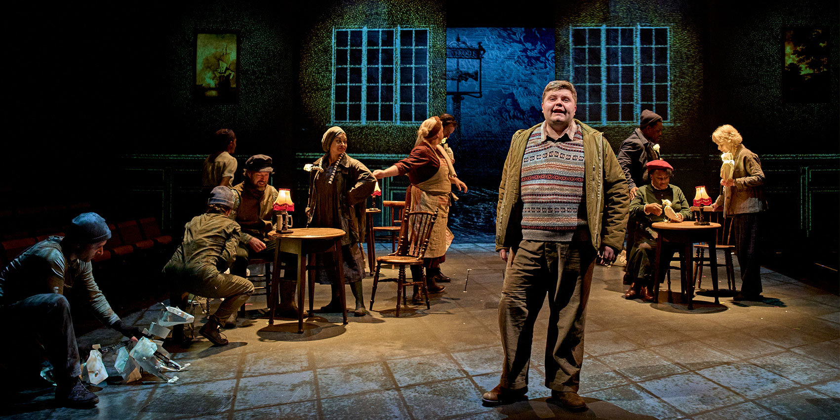 An actor in costume stands under a spotlight with other actors sitting around old-fashioned tables and chairs in the background.
