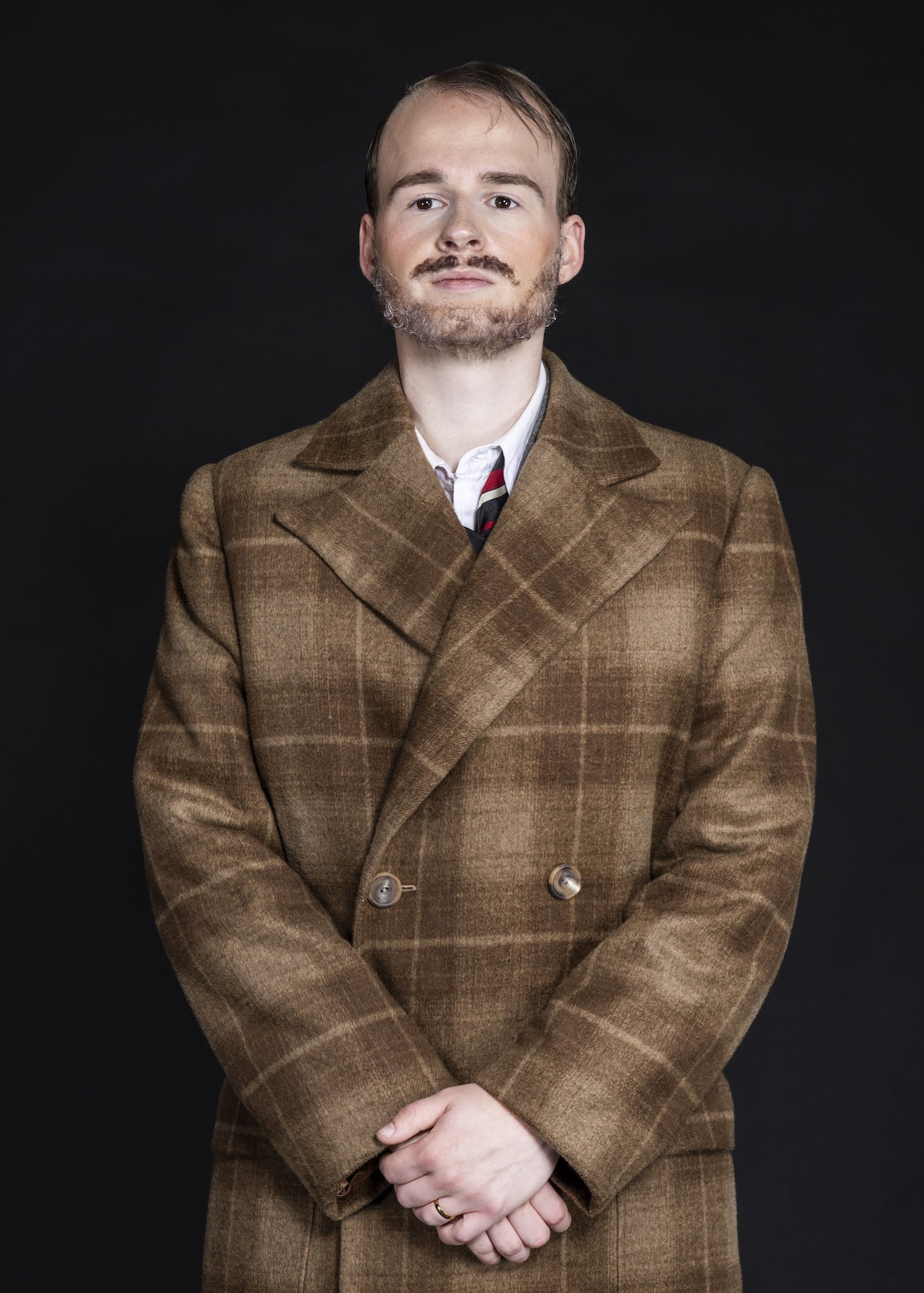 Man with beard wearing brown checked double-breasted coat, white shirt and striped tie, hands clasped, against black background.