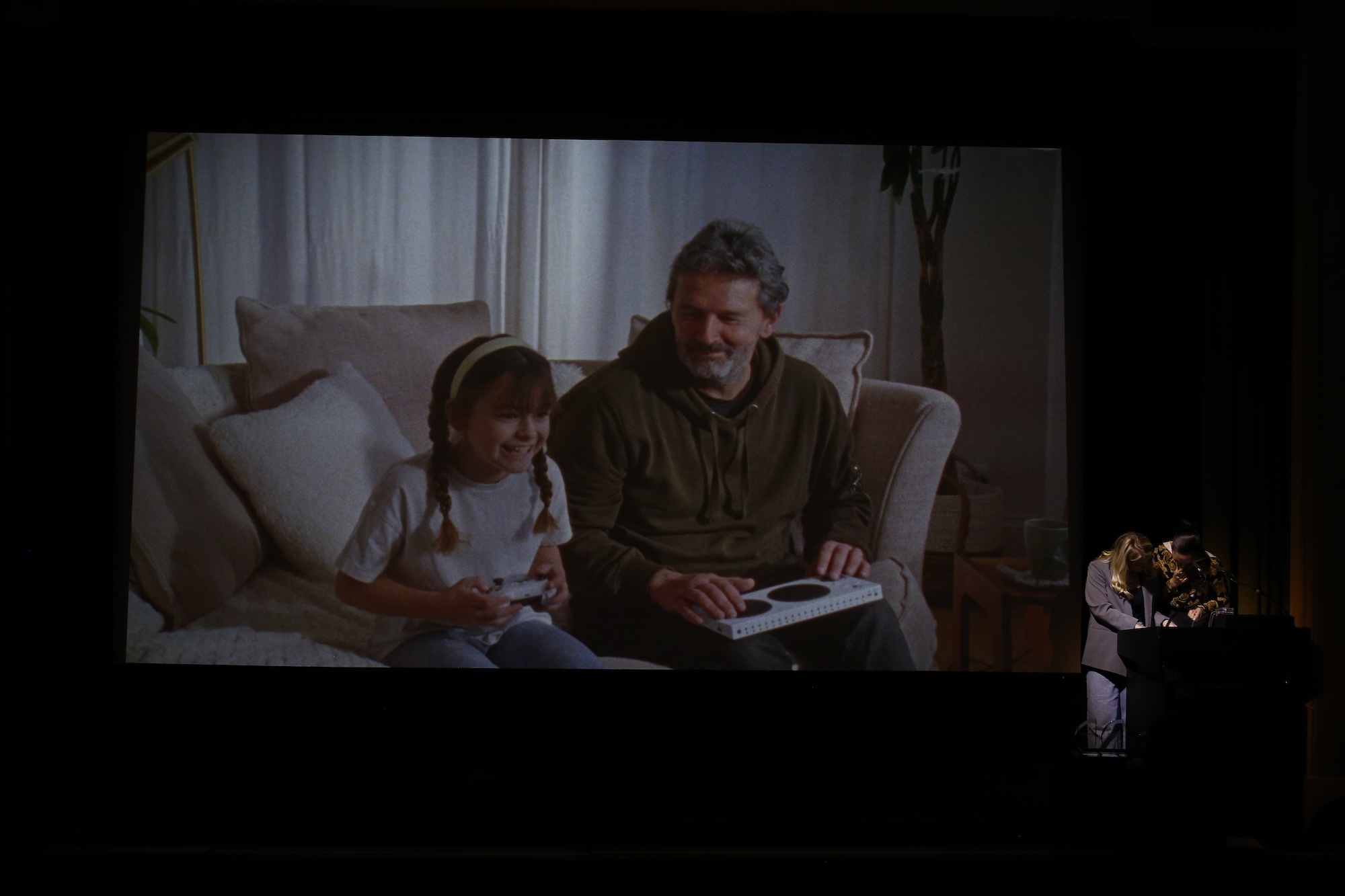 Large screen showing an image from a film, a small girl and her father sitting on a sofa and playing an Xbox.
