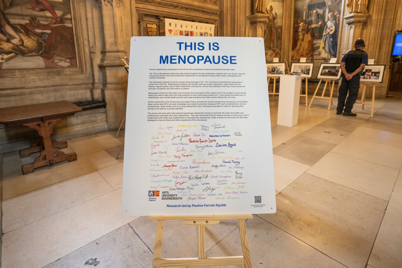 White poster on easel that reads 'this is menopause' in blue, with text explaining below, followed by an array of colourful signatures. Tiled floor and artwork in background.