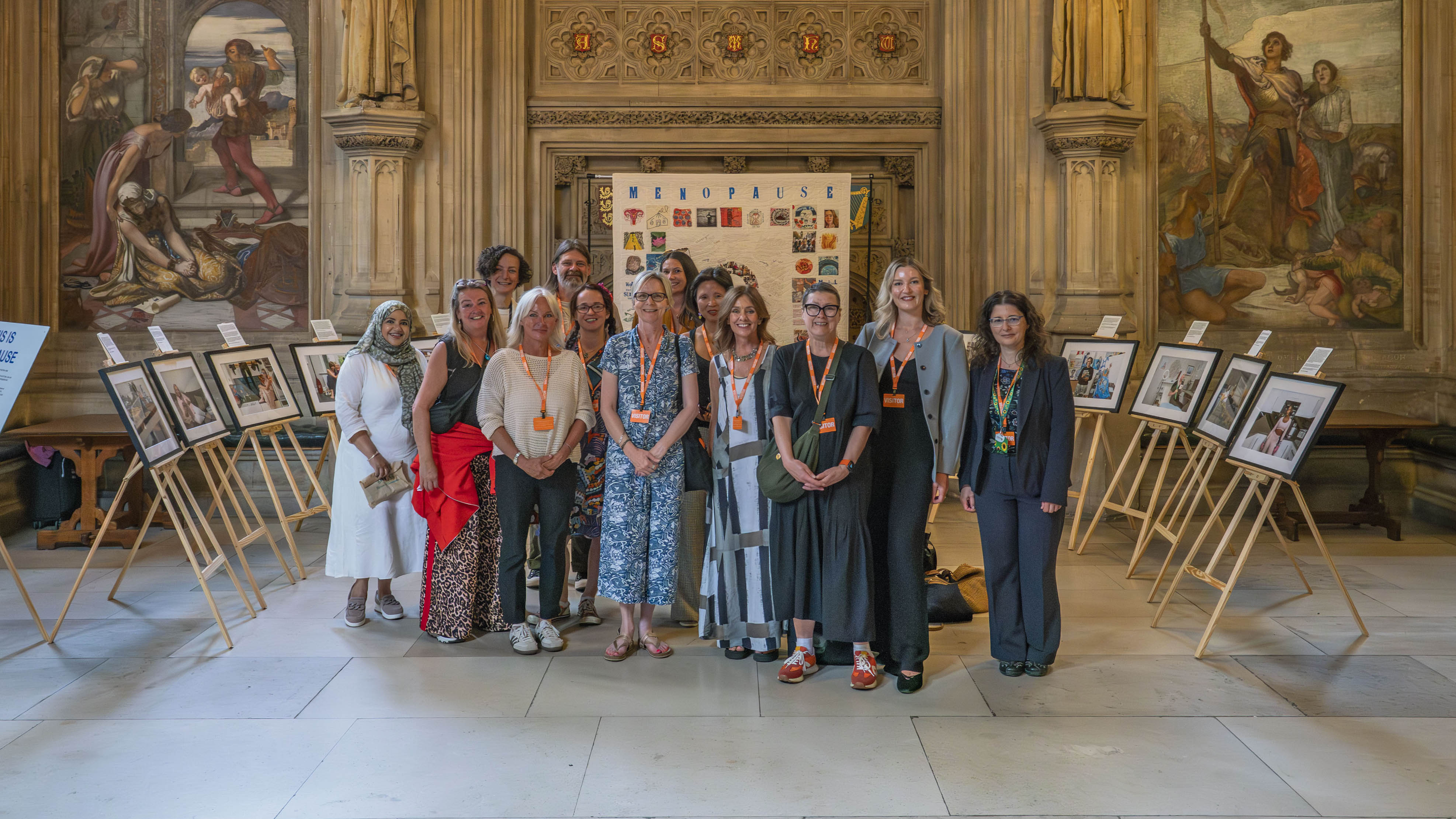 Group of women stood in front of white sheet with 'menopause' title and colourful squares, framed work on easels around them. They are wearing orange lanyards. Gold stone pillars and illustrations behind them.