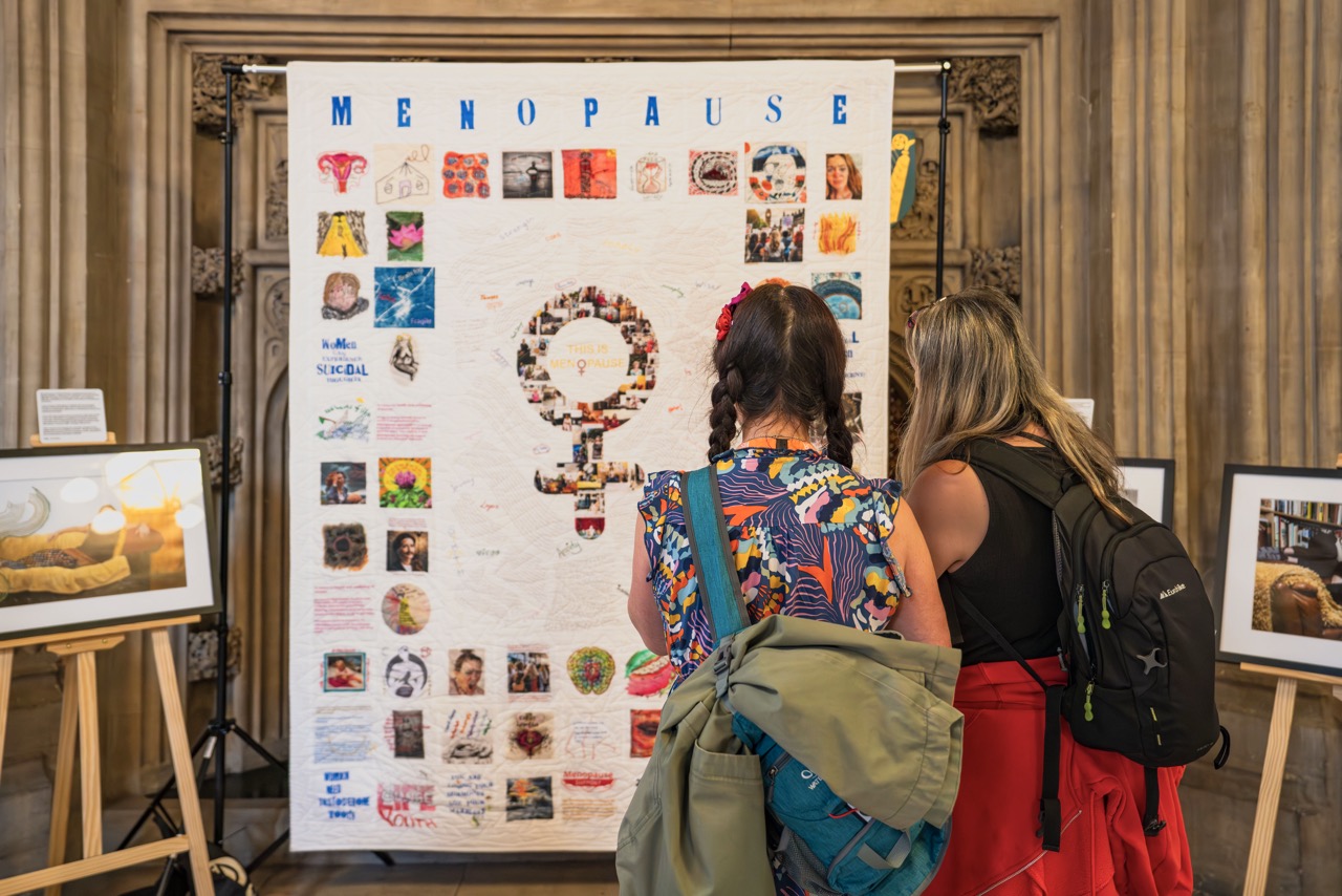 Two people looking at a quilt with colourful squares, showing the female gender symbol in the middle with 'Menopause' in blue above, frames surrounding it.