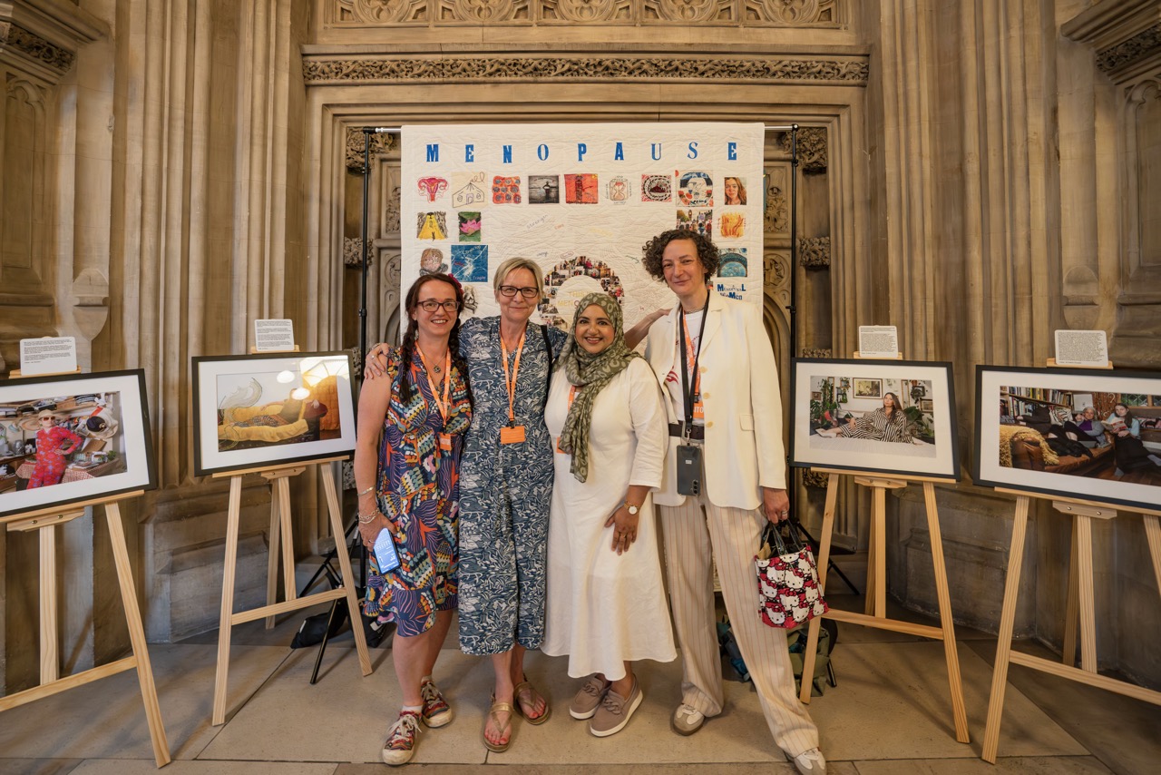 Four women stood smiling in front of the menopause quilt with colourful quilt tiles on a white background, framed artworks on easels surrounding them.