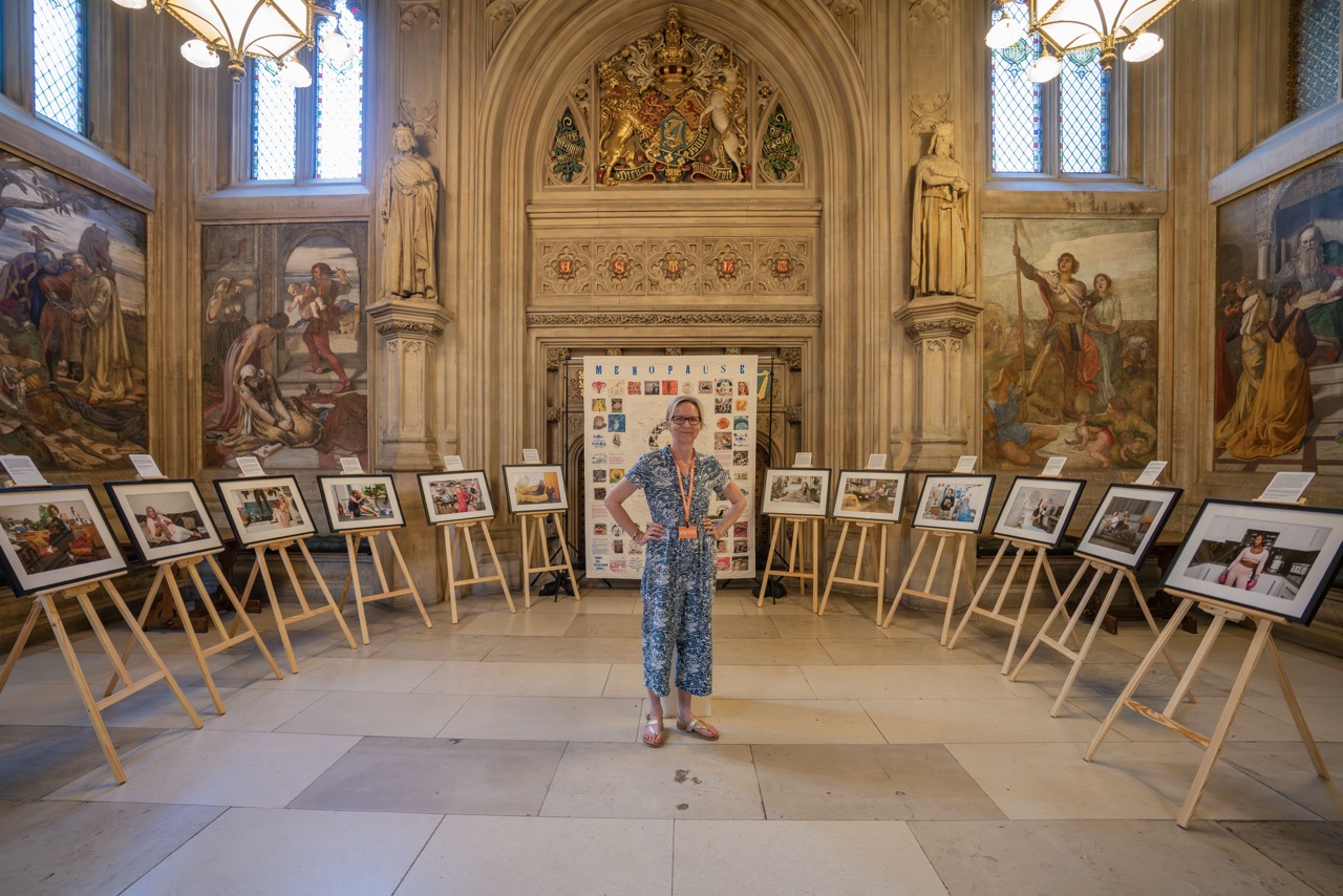 Pauline Ferrick-Squibb stood at the centre of the exhibition, surrounded by frames on easels. She's wearing a blue jumpsuit, detailed illustrations, sculptures and arched door/windows behind her.