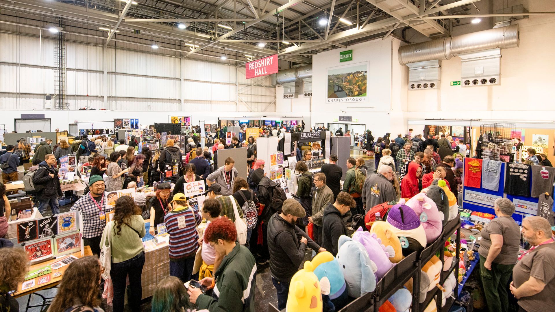 Busy indoor market or trade show with crowds of people browsing vendor stalls in a large warehouse-style venue with exposed ceiling.